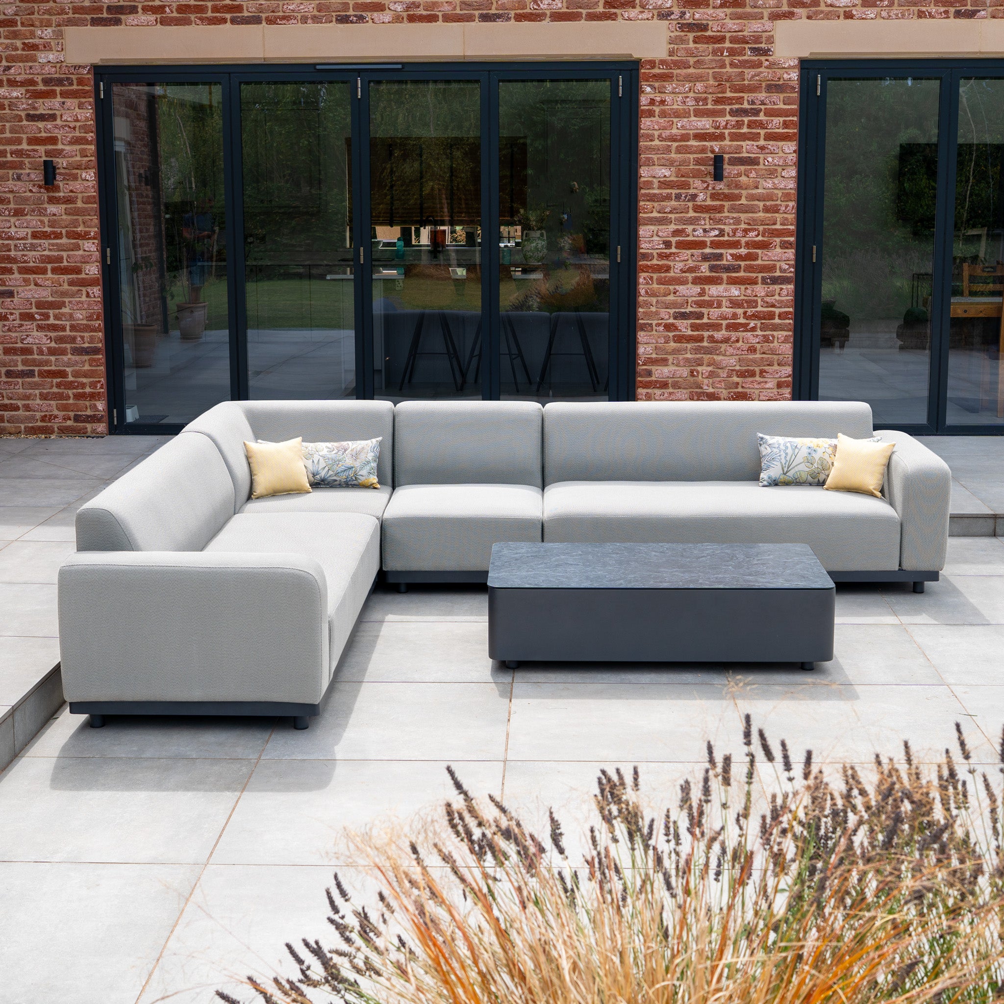 The Luna Outdoor Fabric Large Corner Group Set with Coffee Table in Oyster Grey offers elegant, stain-proof, all-weather garden seating—perfect for patios with its cushions and stylish coffee table in front of glass doors.