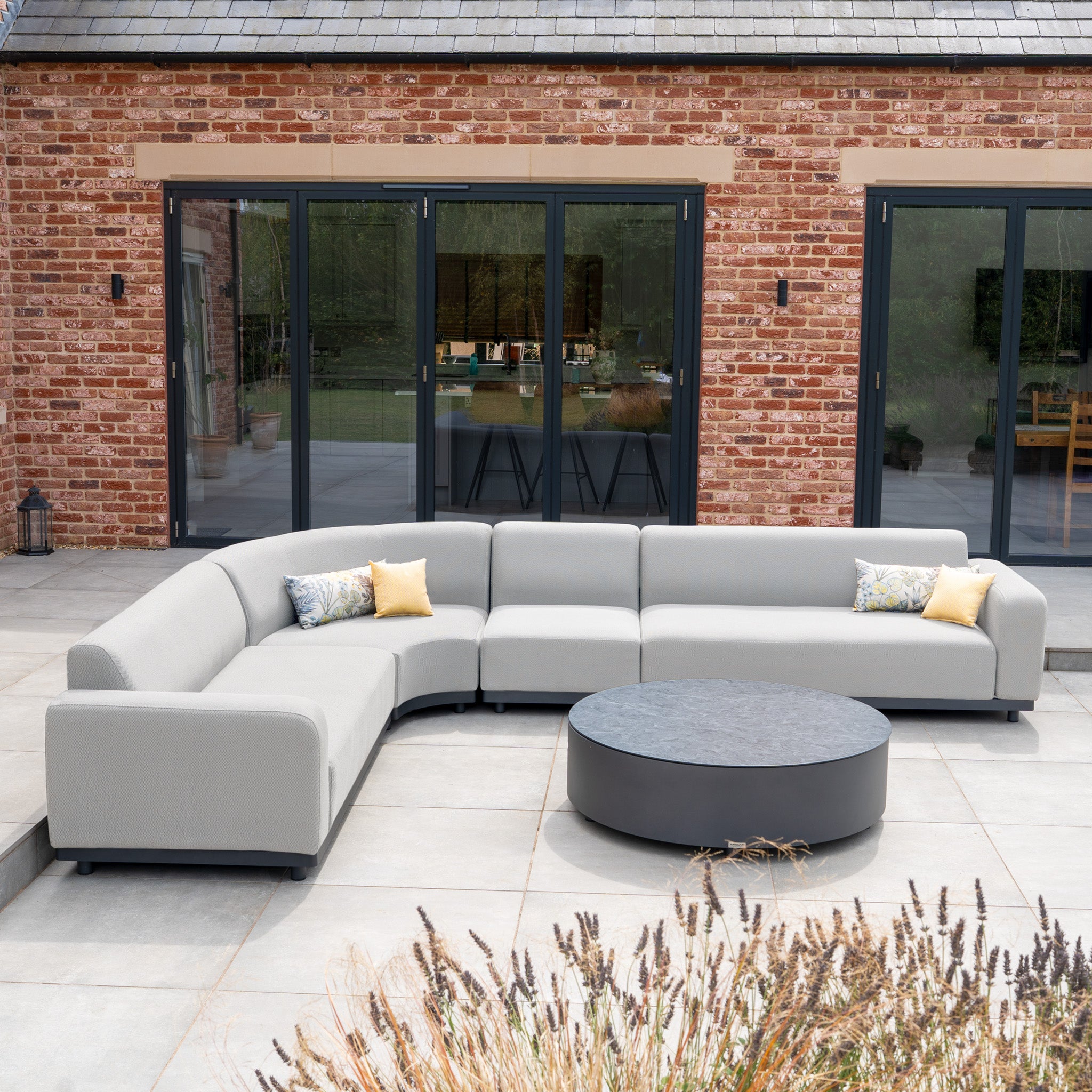 The Luna Outdoor Fabric Large Curved Corner Group Set with Round Coffee Table in Oyster Grey adds elegant, all-weather style to a patio in front of a brick house.