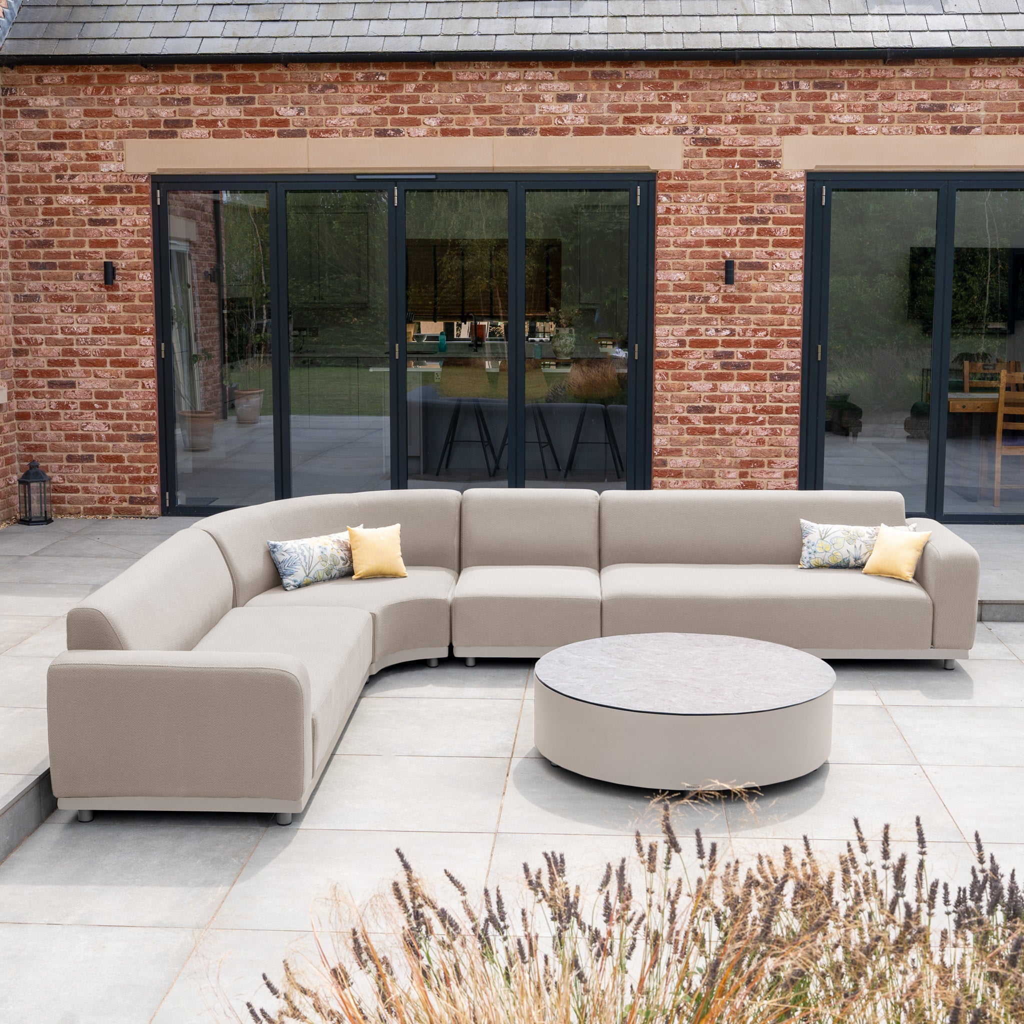 Outdoor patio with a Luna Outdoor Fabric Large Curved Corner Group Set in Fawn and round coffee table, featuring an aluminium frame and yellow accent pillows, placed in front of glass doors.