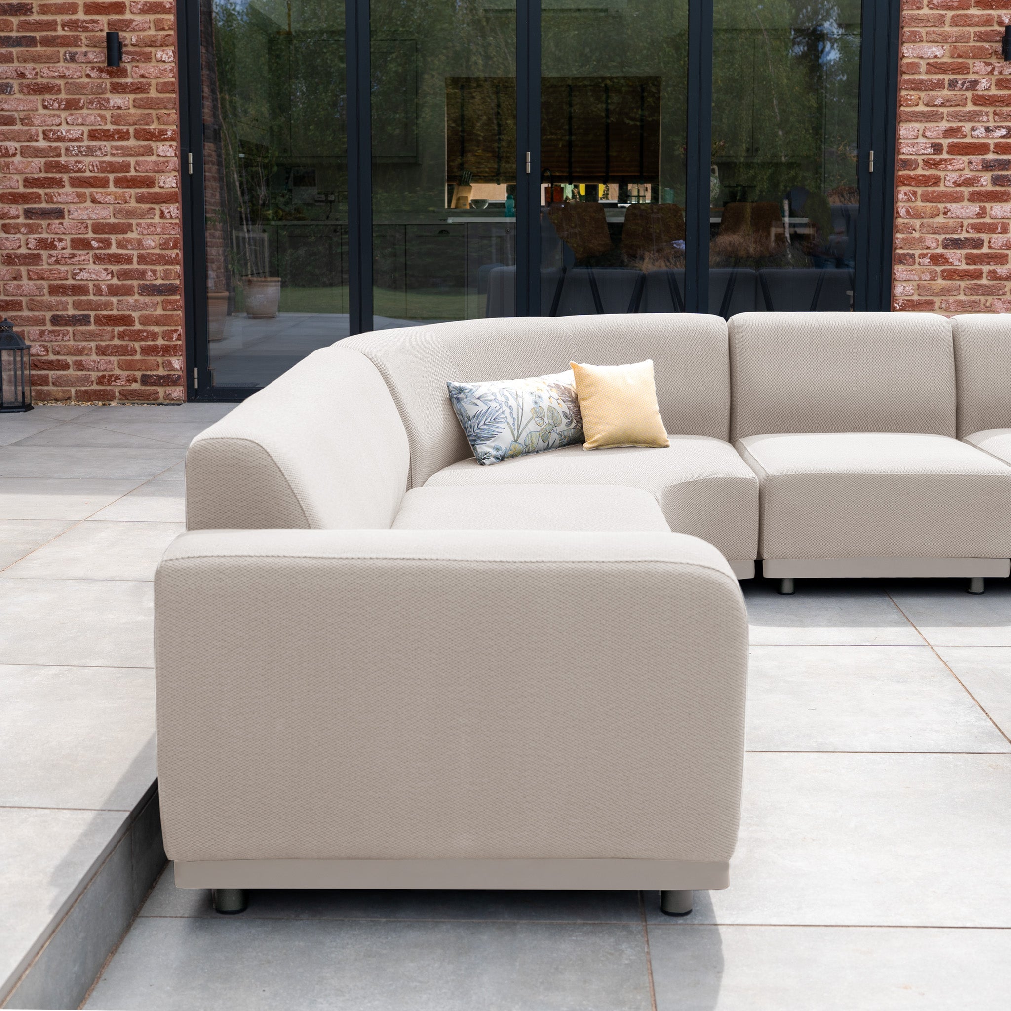 Luna Outdoor Fabric Large Curved Corner Group Set with Round Coffee Table in Fawn features beige all-weather cushions, two decorative pillows, and is shown on a tiled patio by glass doors and a brick wall.