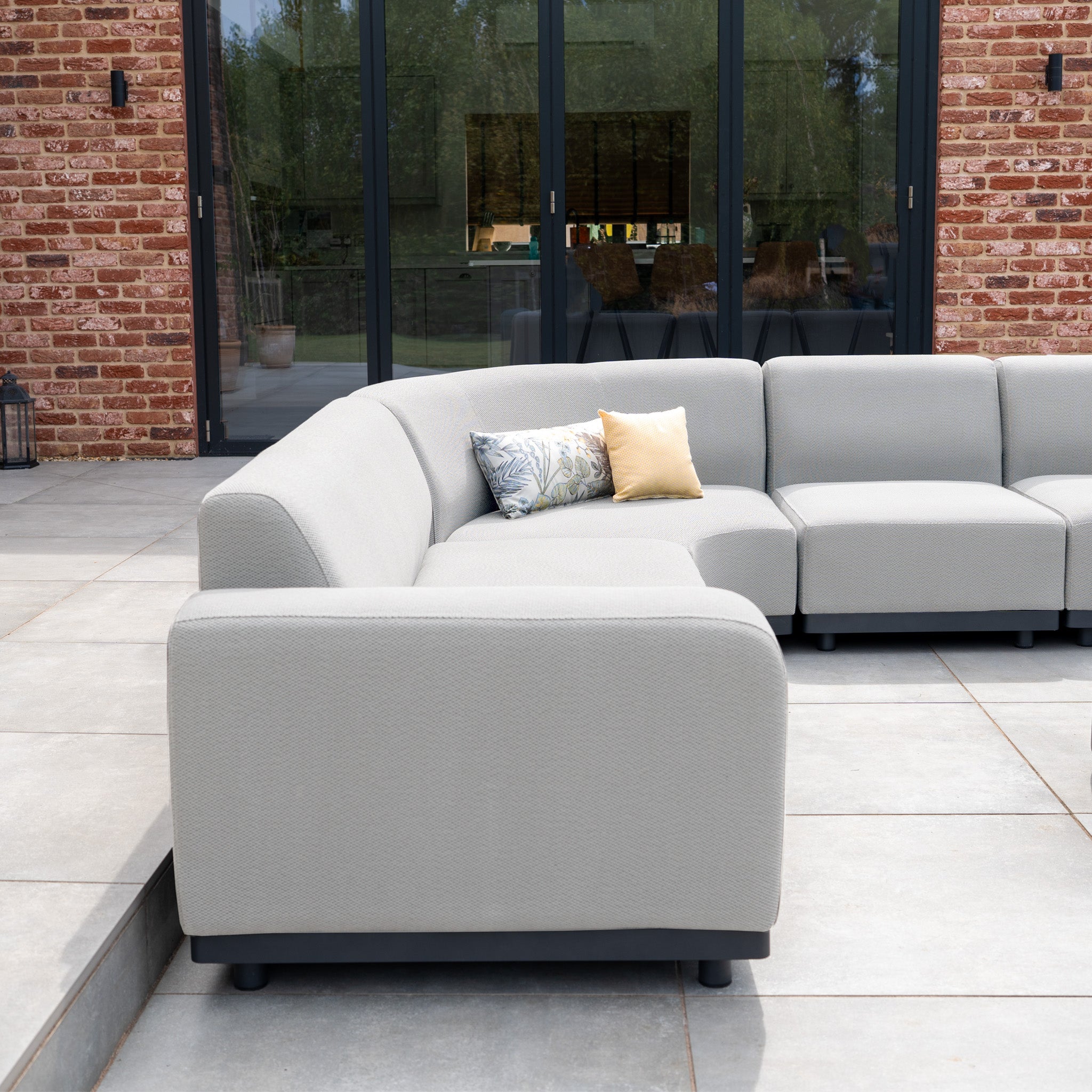 The Luna Outdoor Fabric Large Curved Corner Group Set with Round Coffee Table in Oyster Grey features a weather-resistant sectional and decorative pillows, perfect for patios with glass doors and brick walls.