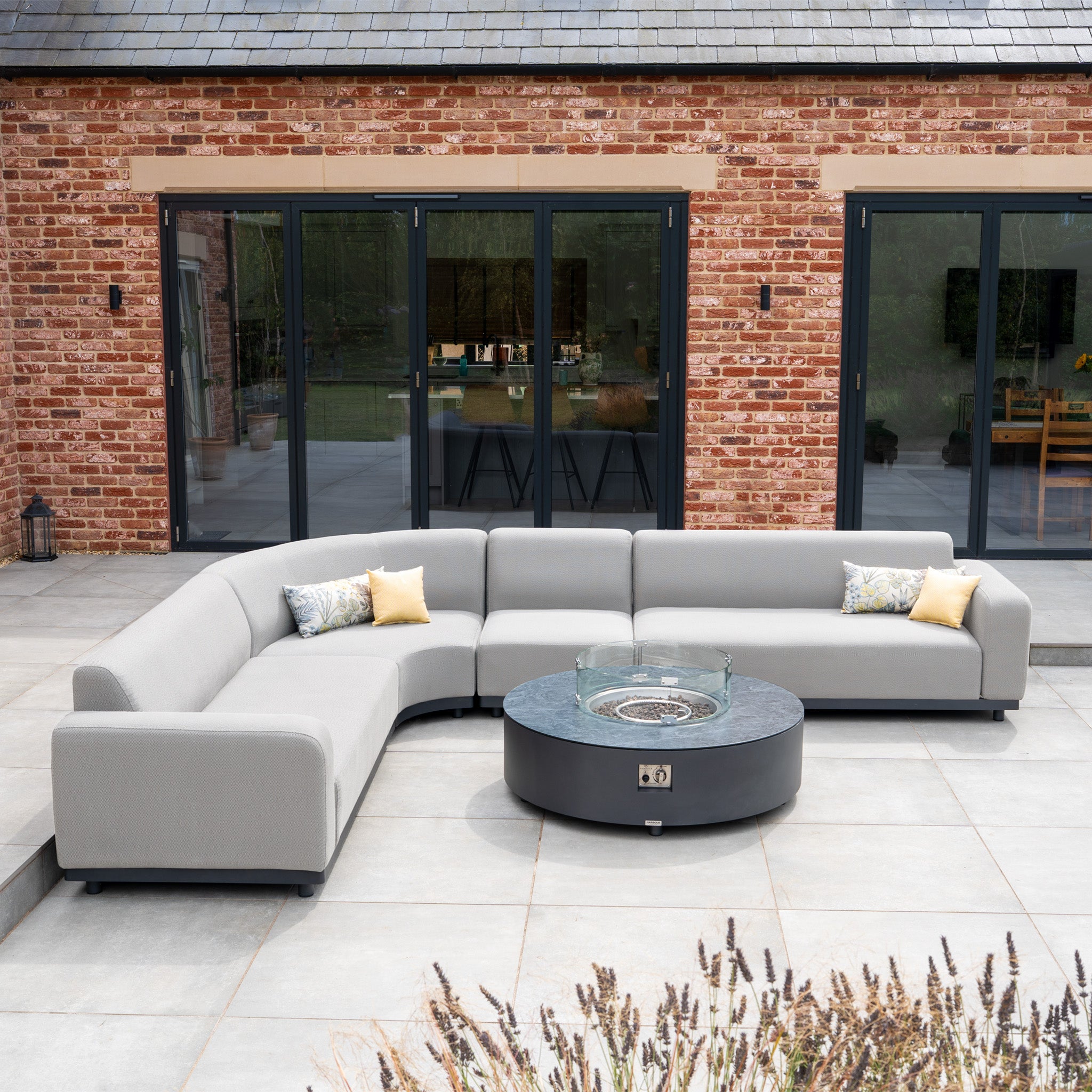 The Luna Outdoor Fabric Large Curved Corner Group Set with Round Firepit Coffee Table in Oyster Grey sits on a patio before glass doors and a brick wall.