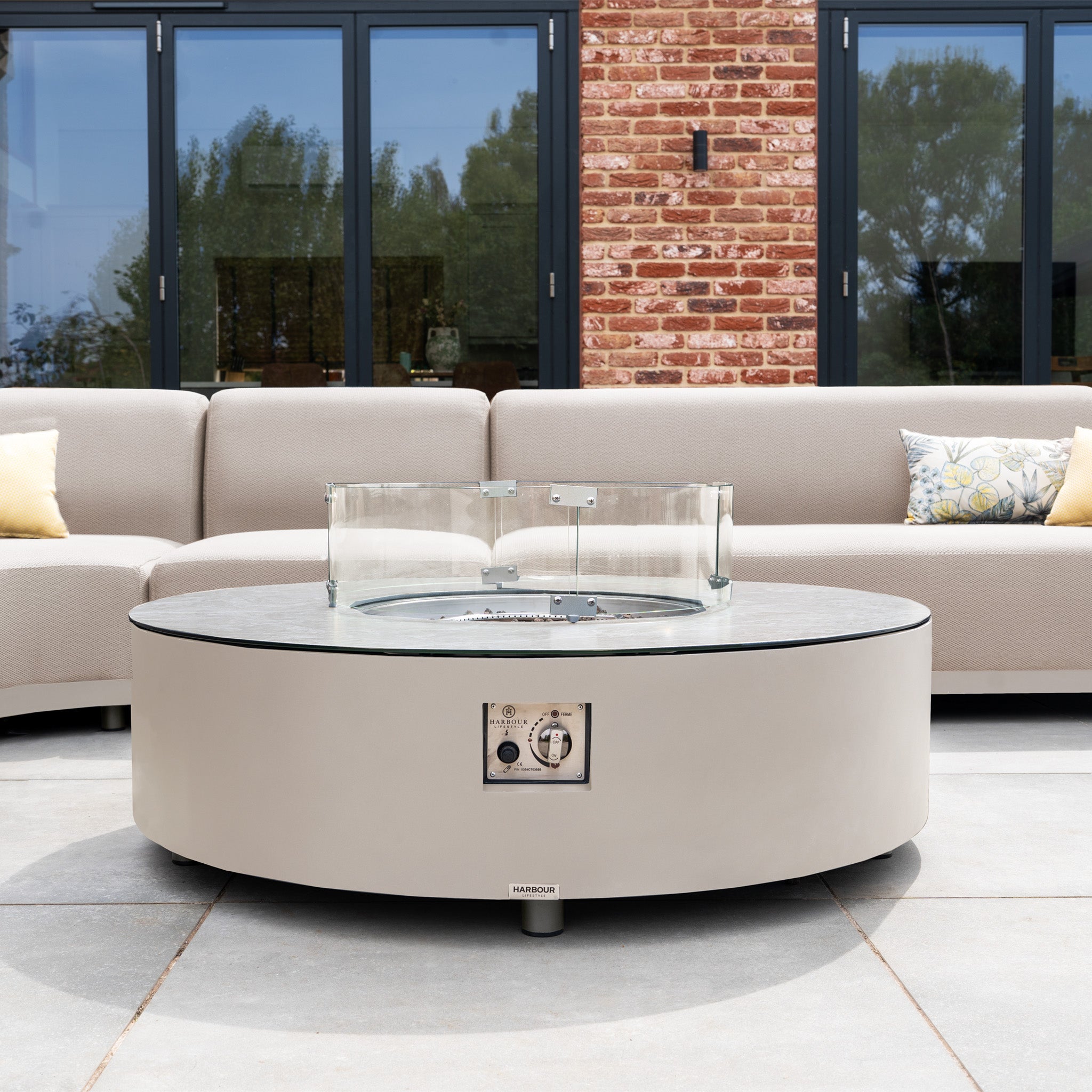The Luna Outdoor Fabric Large Curved Corner Group Set with Round Firepit Coffee Table in Fawn is arranged on the patio, surrounded by beige all-weather garden furniture and cozy cushions.