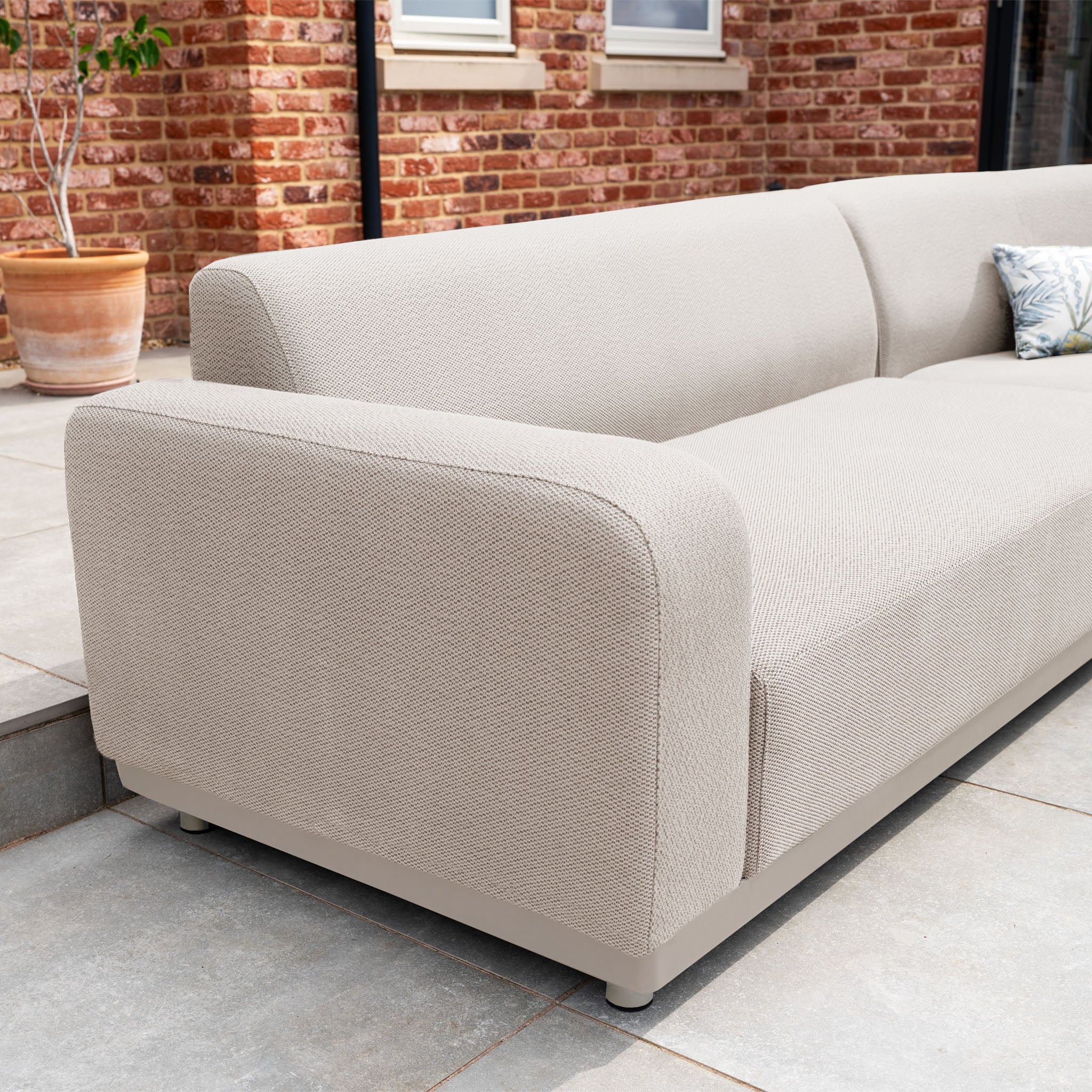 The Luna Outdoor Fabric Large Curved Corner Group Set with Round Firepit Coffee Table in Fawn is shown on a tiled patio, set against a brick wall with a potted plant in the background.