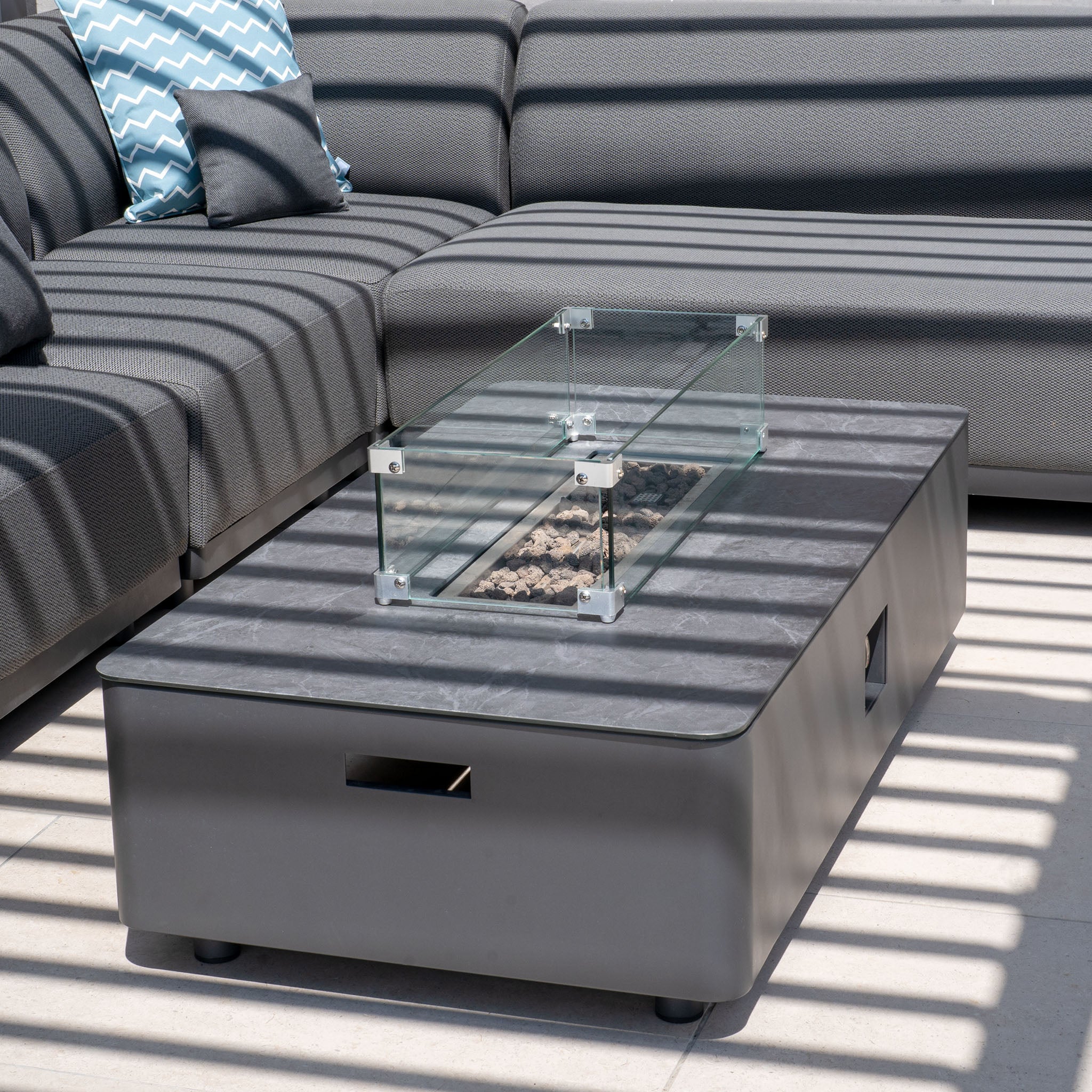 Outdoor seating area showcasing the Luna Outdoor Fabric Large Corner Group Set in Grey, paired with a firepit coffee table, all nestled under striped shadows for an ideal mix of style and comfort.