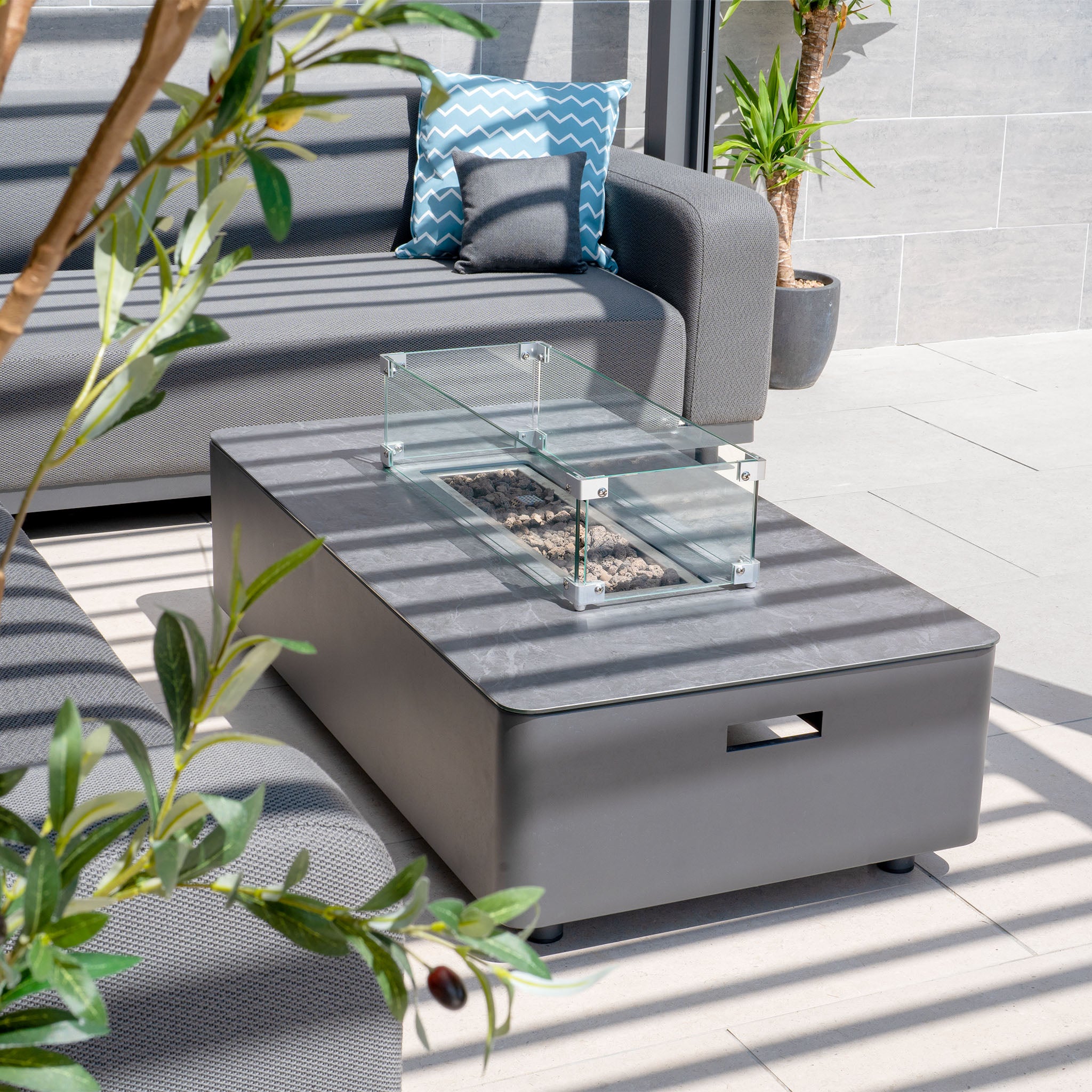 A modern patio displays the Luna Outdoor Fabric Large Corner Group Set in Grey with a firepit coffee table, highlighted by a blue patterned pillow and leafy plant for stylish durability.