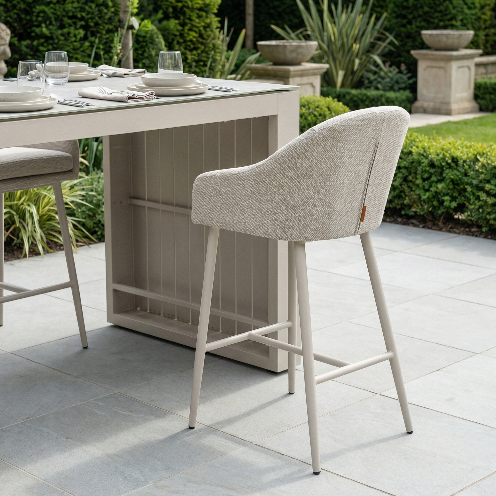 The Luna Outdoor Fabric Bar Stool in Fawn sits at a set dining table on a tiled patio, featuring weather-resistant cushions and overlooking a lush green garden.