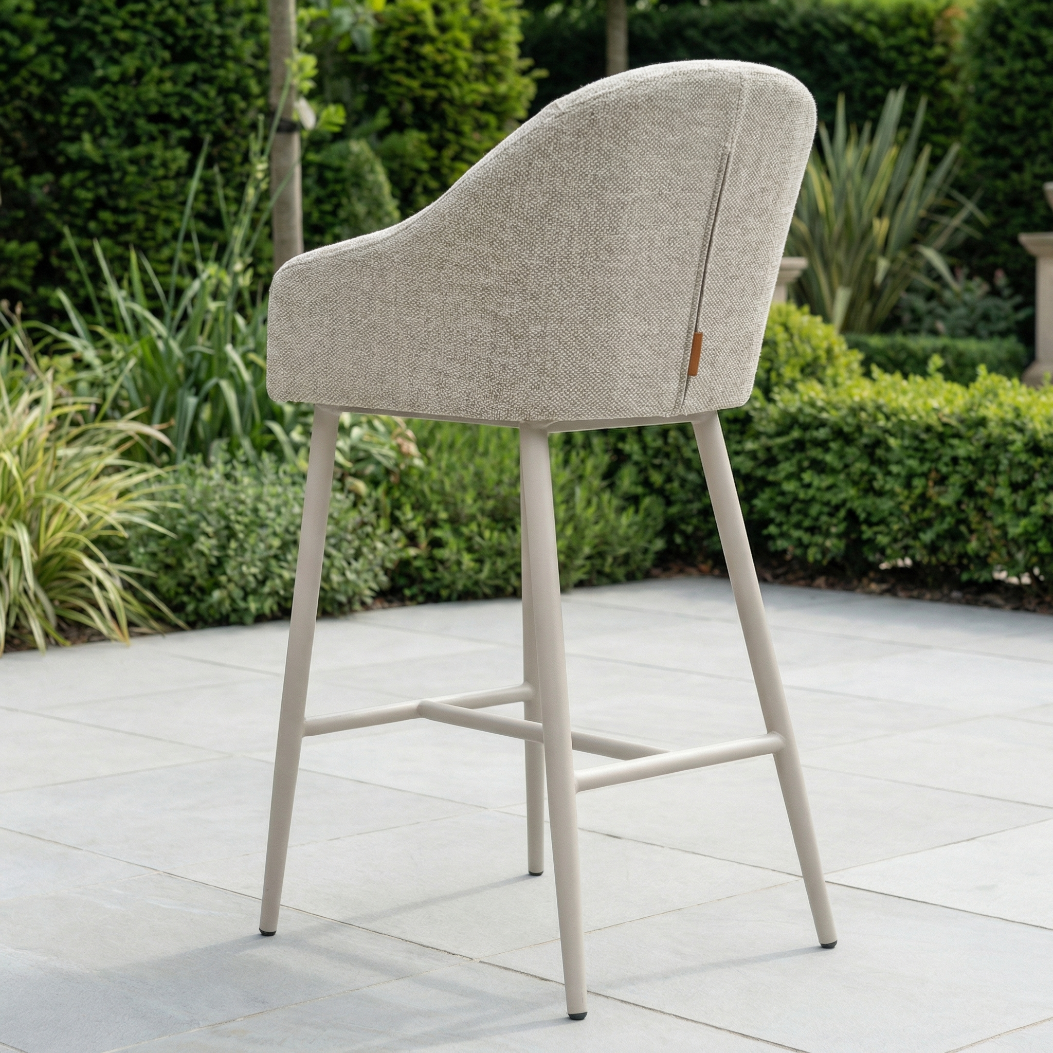 The Luna Outdoor Fabric Bar Stool in Fawn, featuring weather-resistant cushions and metal legs, is placed on a stone patio amid a landscaped garden.