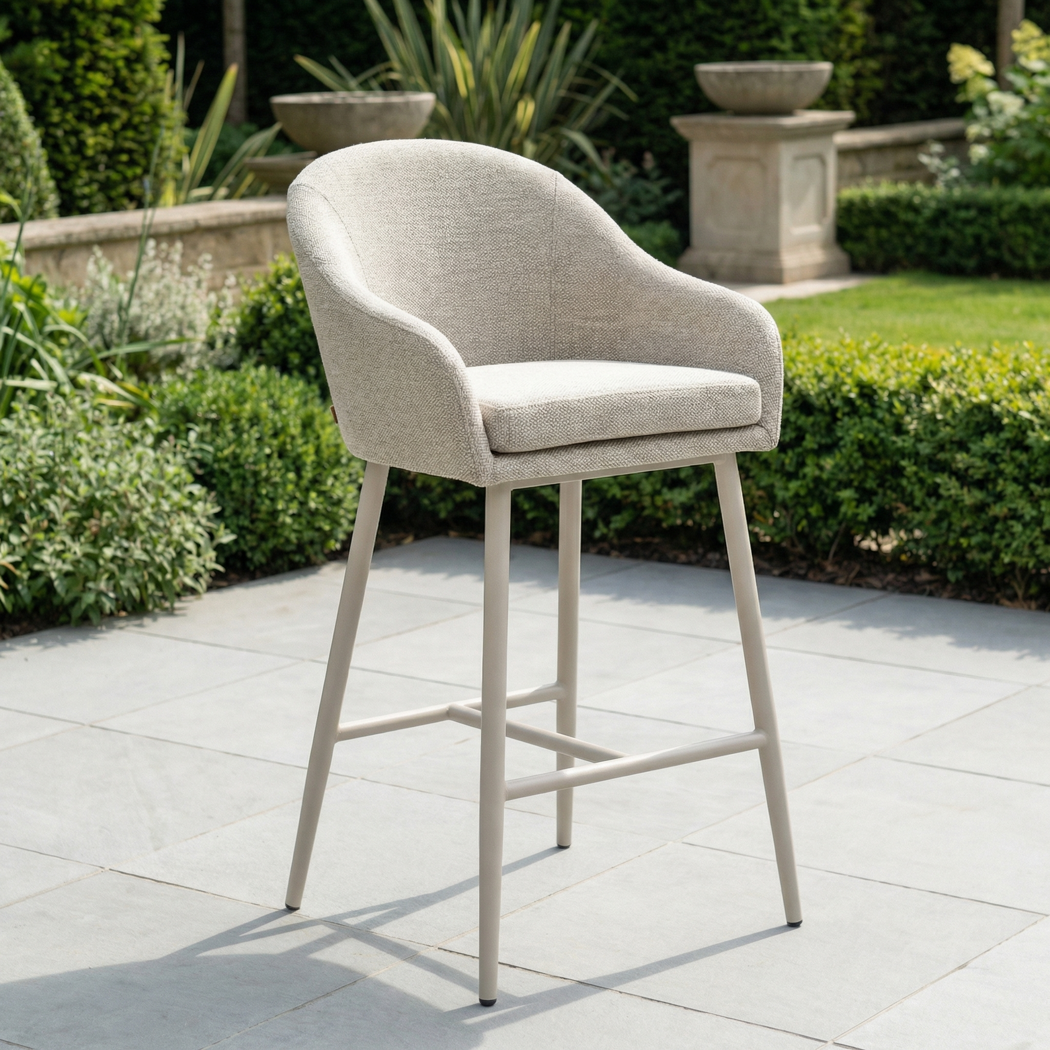 The Luna Outdoor Fabric Bar Stool in Fawn features slim metal legs and weather-resistant cushions, ideal for stylish outdoor comfort on a stone patio surrounded by lush garden greenery.