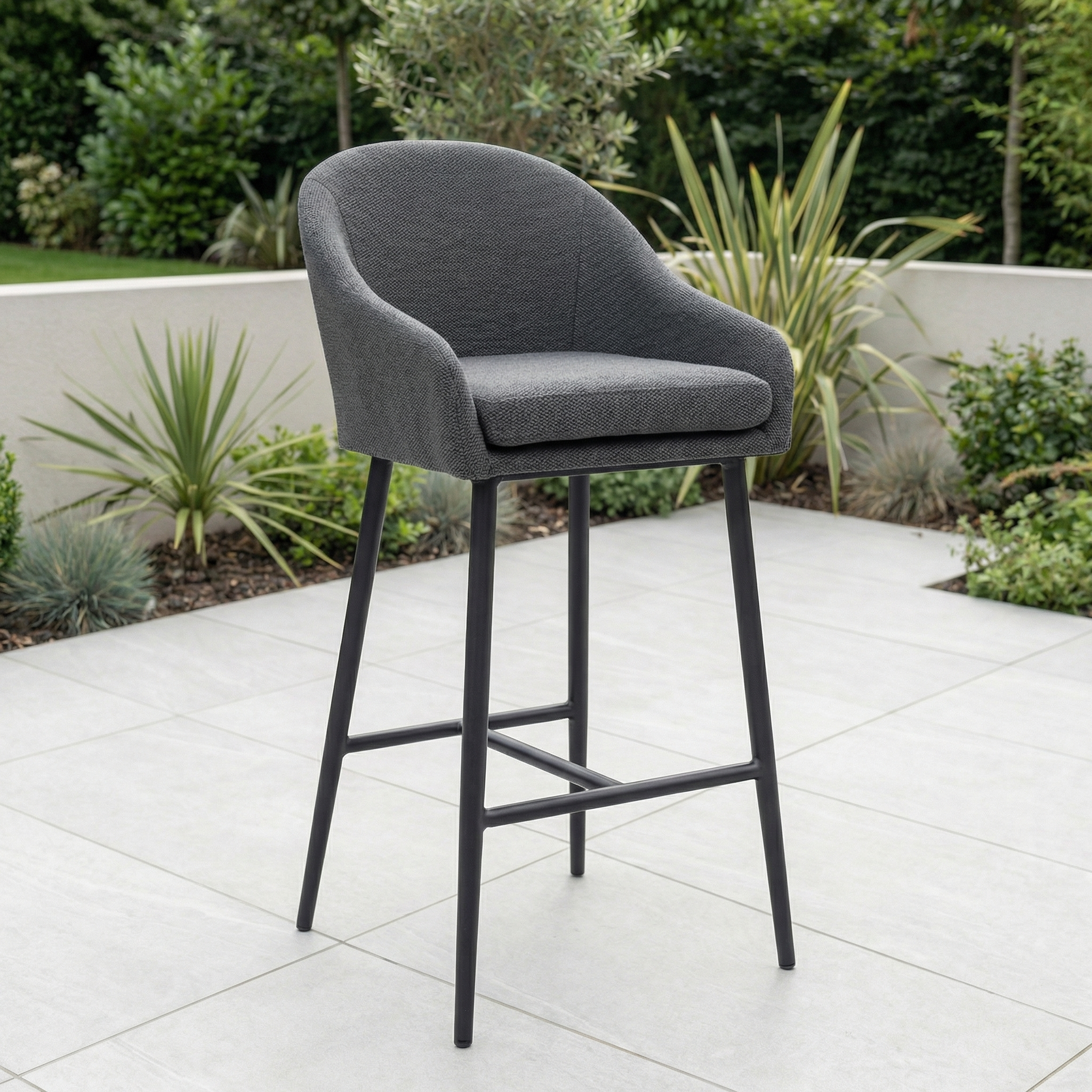 The Luna Outdoor Fabric Bar Stool in Grey, with armrests and weather-resistant cushions, stands on a tiled patio surrounded by lush green plants and shrubs.