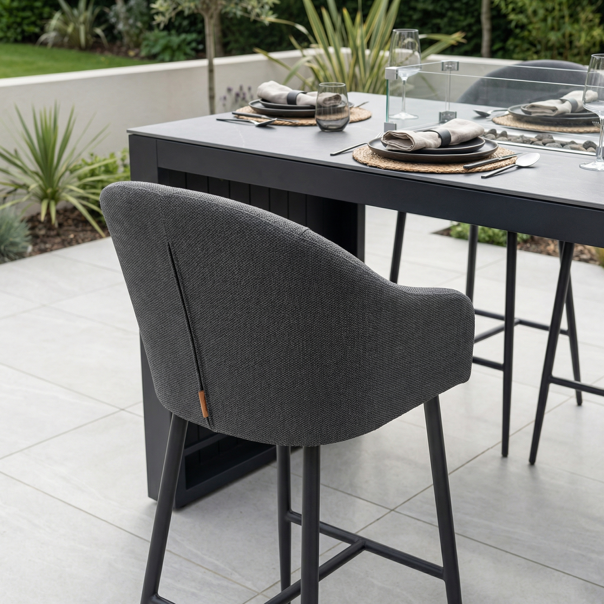 Modern outdoor dining area with the Luna Outdoor Fabric Bar Stool in Grey, featuring weather-resistant cushions and a set table on a tiled patio surrounded by plants.