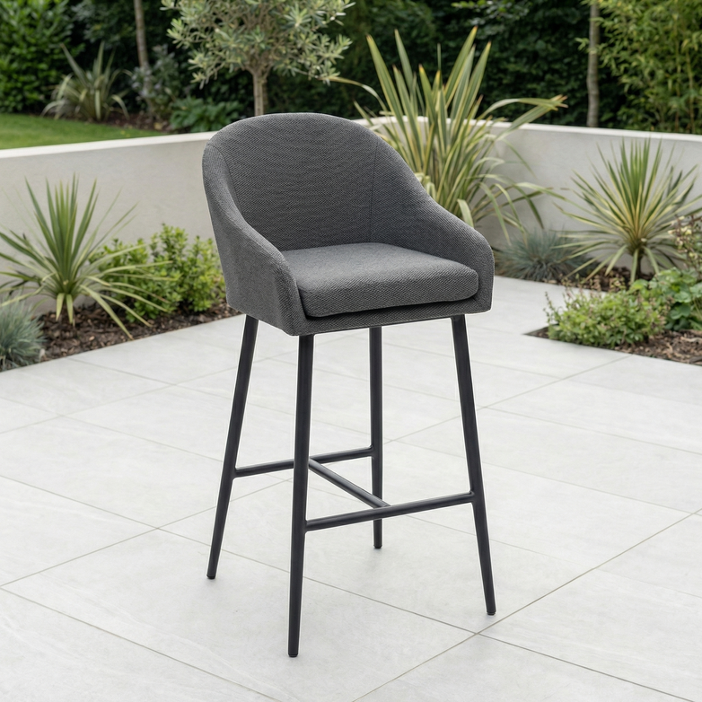 The Luna Outdoor Fabric Bar Stool in Grey features metal legs and weather-resistant cushions, perfect for lasting comfort on a tiled patio surrounded by garden plants.