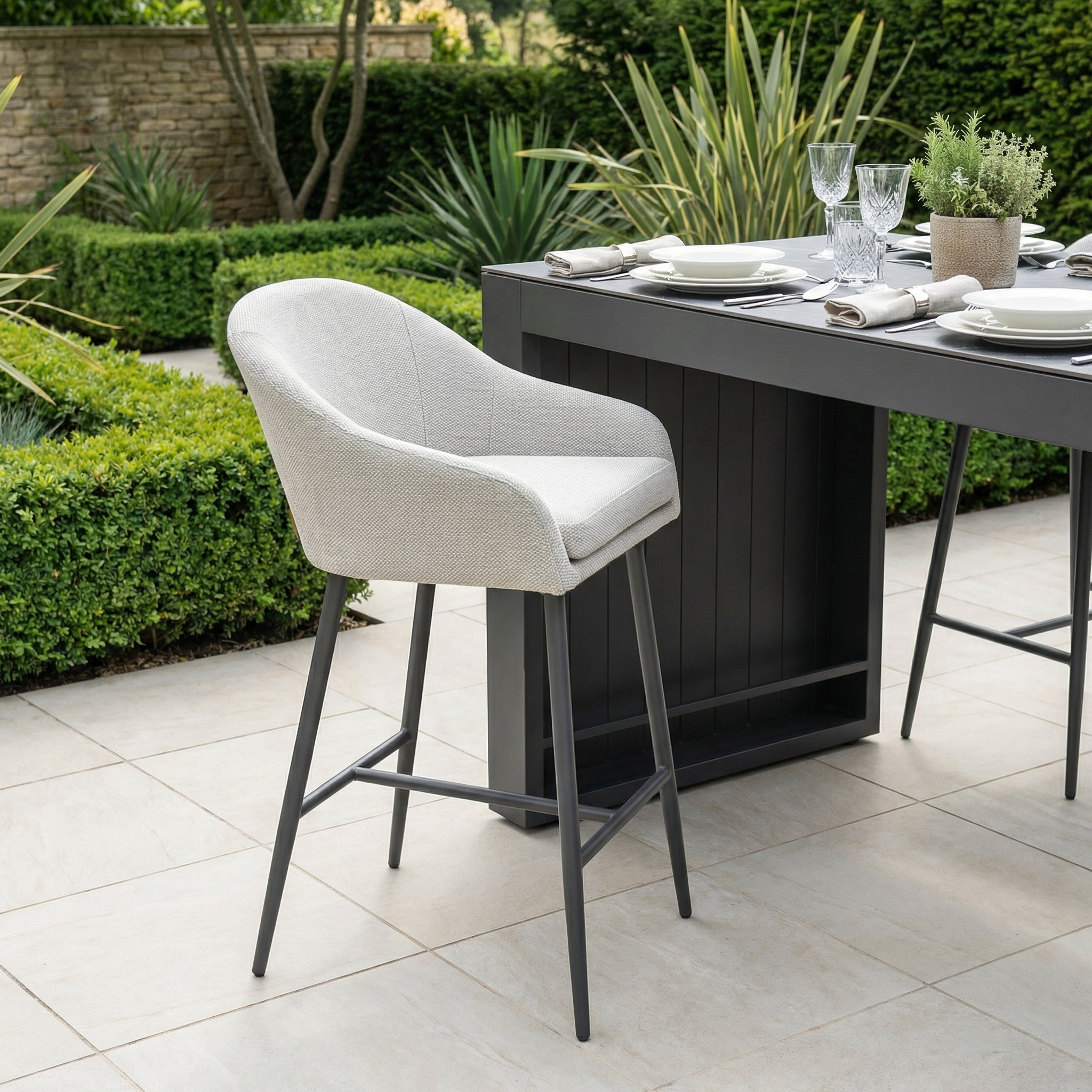A modern outdoor dining area featuring the Luna Outdoor Fabric Bar Stool in Oyster Grey with weather-resistant cushions and a black table set for a meal.