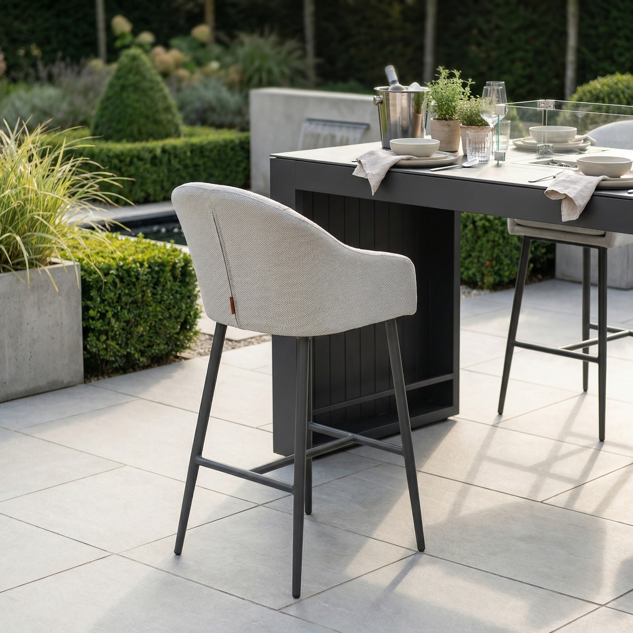 Modern outdoor dining area featuring Luna Outdoor Fabric Bar Stools in Oyster Grey with weather-resistant cushions around a table set for a meal on a tiled patio, surrounded by lush greenery.