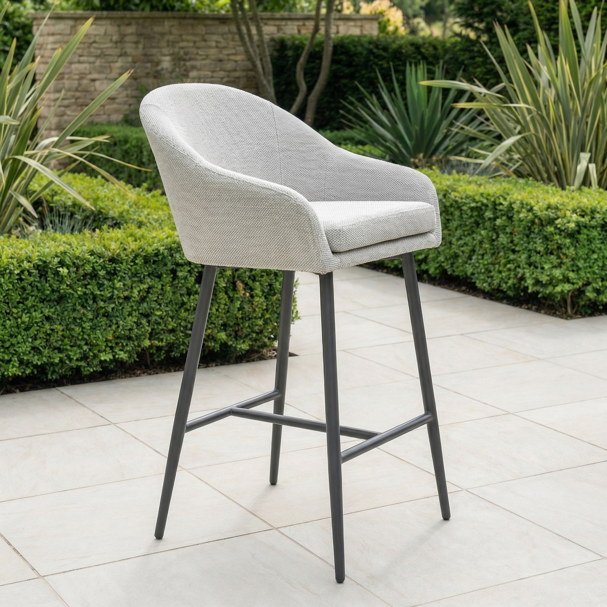 The Luna Outdoor Fabric Bar Stool in Oyster Grey features cushioned seating and black legs, perfect for a tiled patio. Weather-resistant cushions provide lasting comfort and durability, ideal for use among green bushes and plants outdoors.