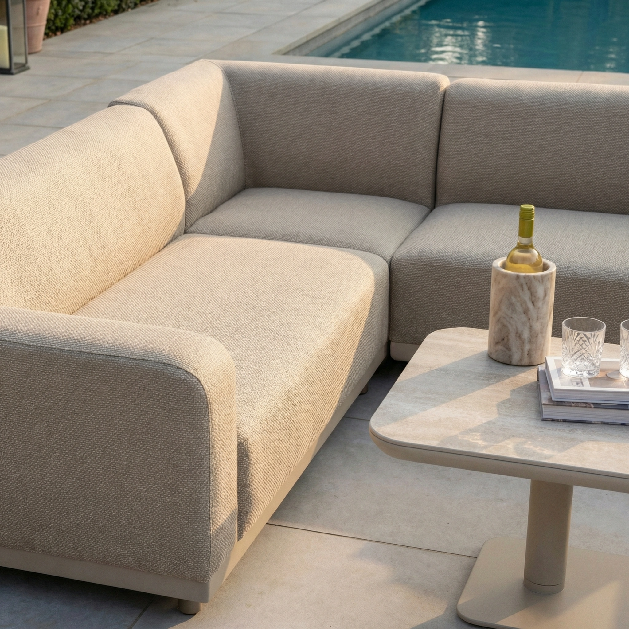 The Luna Outdoor Fabric Corner Sofa Set with Altura Rising Table in Fawn, crafted from stain-resistant, all-weather material, sits poolside with wine, glasses, and books basking in the sunlight on the table.