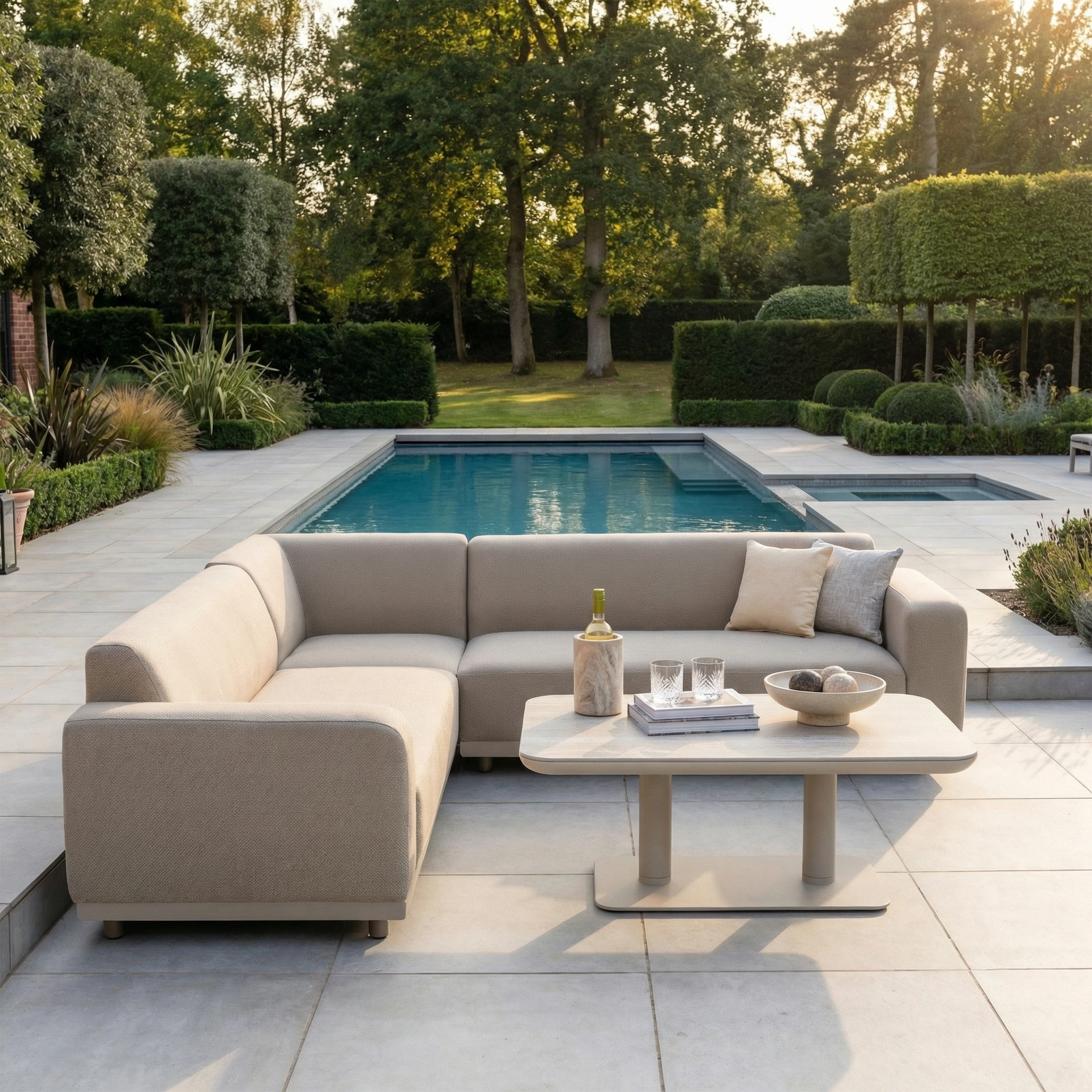 The Luna Outdoor Fabric Corner Sofa Set with Altura Rising Table in Fawn sits by the pool, surrounded by greenery. Made with all-weather materials and stain-resistant fabric, it's a stylish, durable choice for any season.