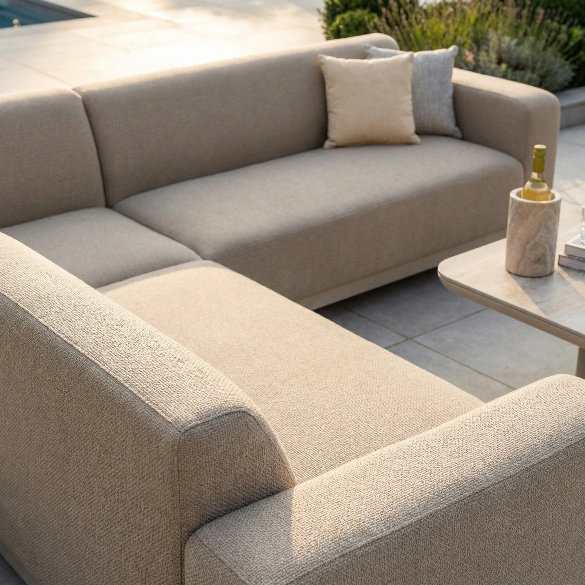 The Luna Outdoor Fabric Corner Sofa Set with Altura Rising Table in Fawn is crafted from stain-resistant, all-weather material and includes two pillows; its small table holds a wine bottle and a glass.