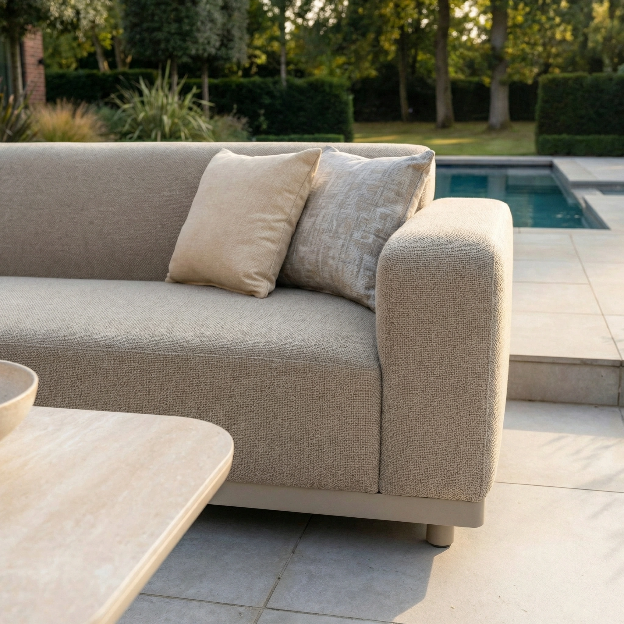 The Luna Outdoor Fabric Corner Sofa Set with Altura Rising Table in Fawn features stain-resistant, all-weather fabric and two pillows, shown on a patio by a pool with trees in the background.