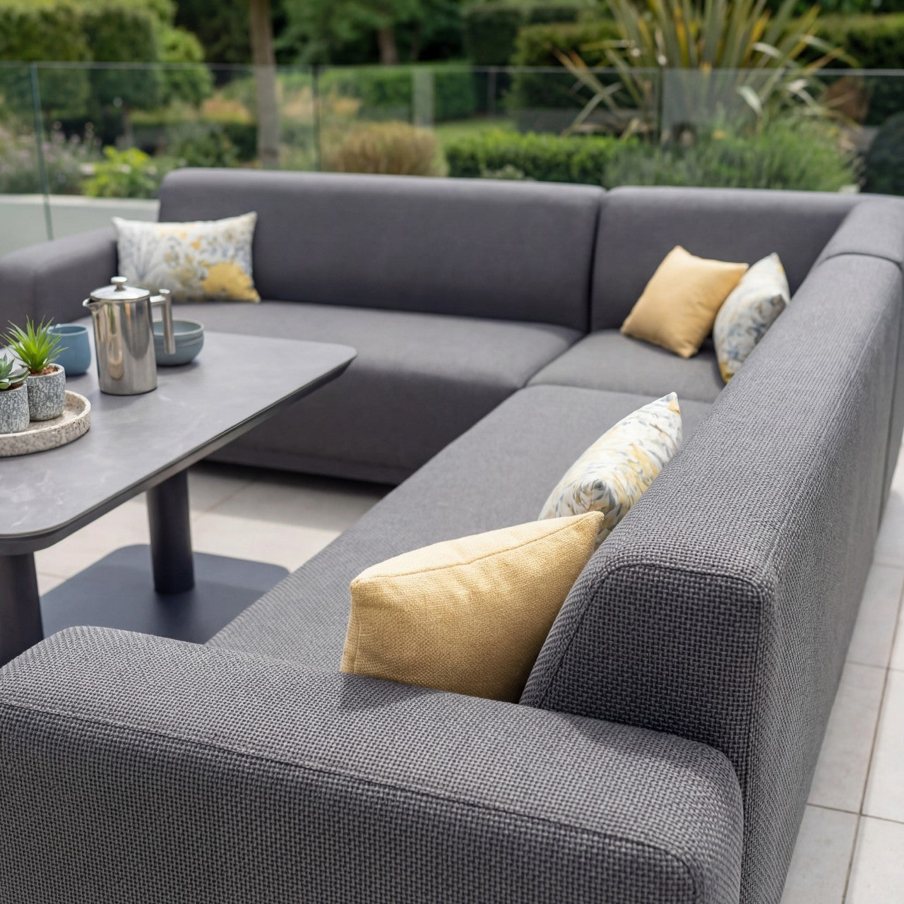 The Luna Outdoor Fabric Corner Sofa Set with Altura Rising Table in Grey features stain-resistant fabric and is accented with yellow and patterned pillows, plus a table for teapot and plants—a stylish garden furniture choice.