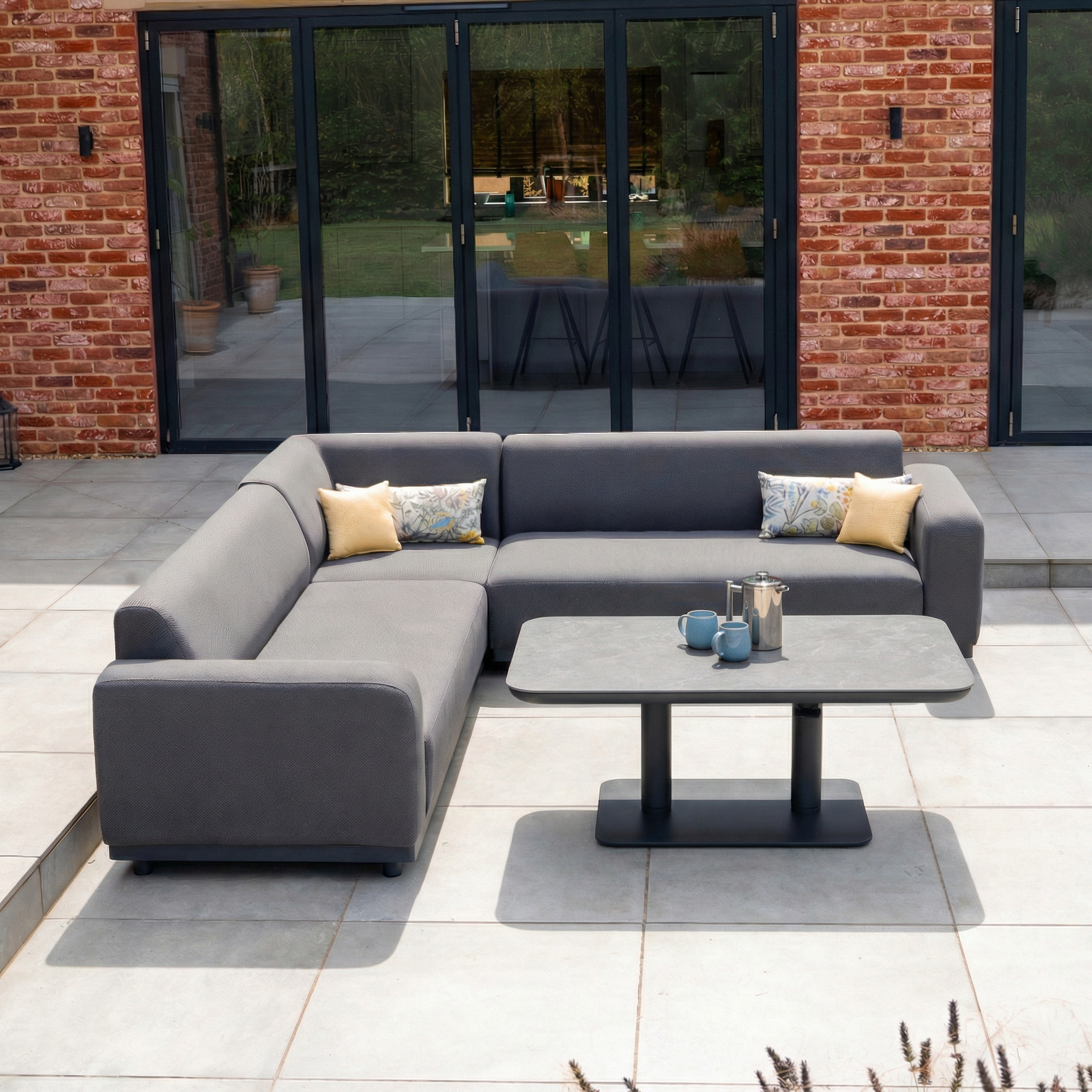 The Luna Outdoor Fabric Corner Sofa Set with Altura Rising Table in Grey, made from stain-resistant fabric, is ideal garden furniture for relaxing on the patio, as seen here against a brick wall and glass doors.