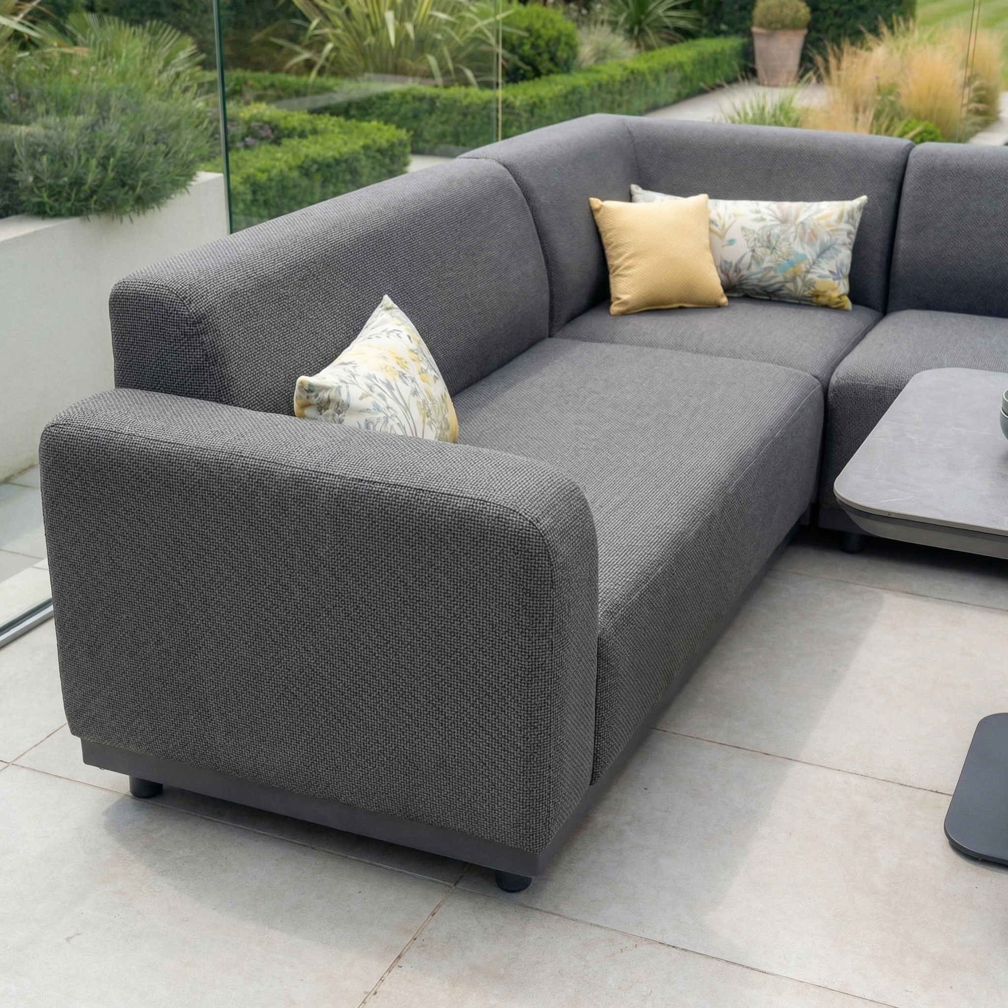 The Luna Outdoor Fabric Corner Sofa Set with Altura Rising Table in Grey features stain-resistant fabric and decorative pillows, making it a stylish and durable garden furniture set for your patio, overlooking a landscaped garden.