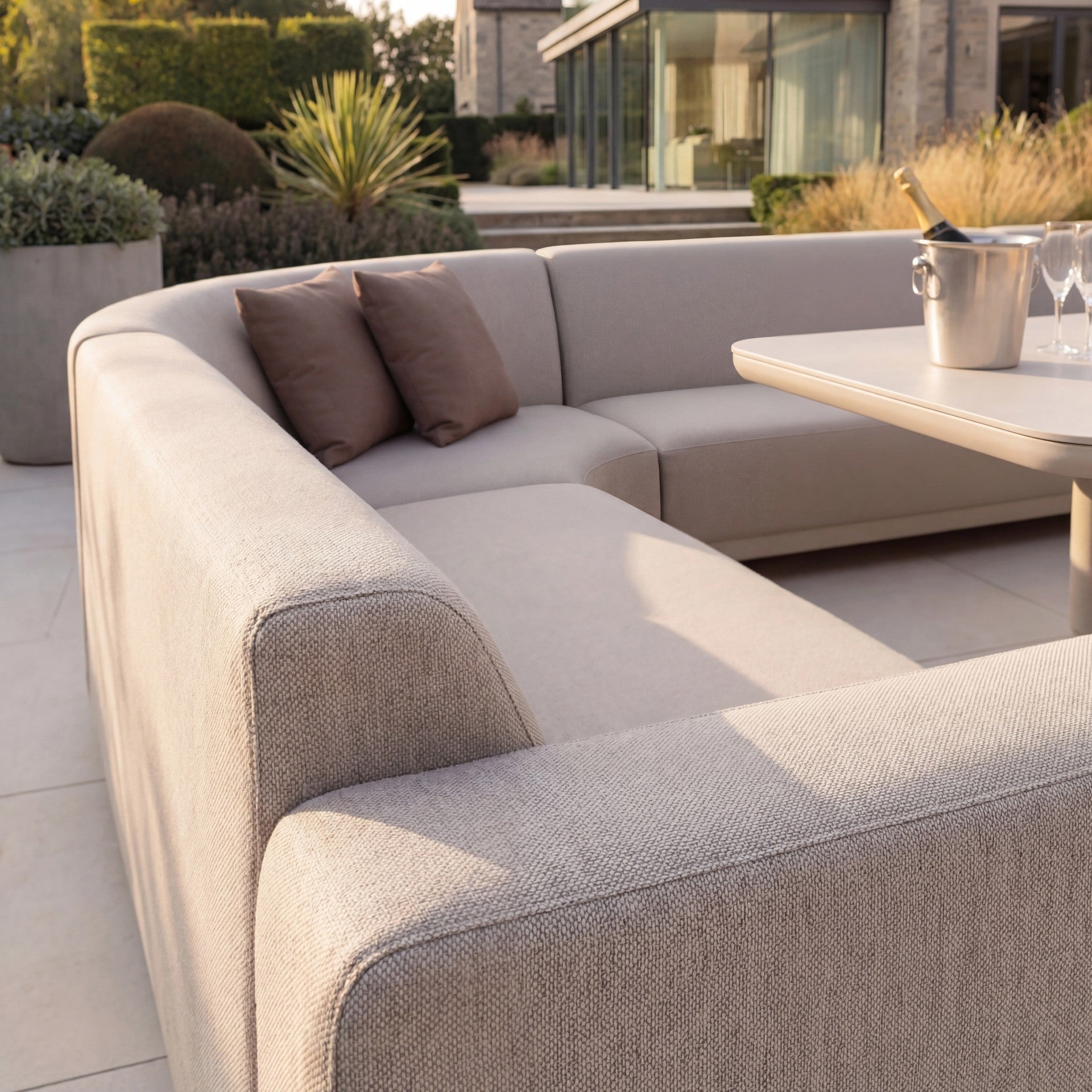 Modern patio featuring the Luna Outdoor Fabric Curved U-Shape Sofa Set in Fawn with two brown pillows and an Altura Rising Table set with champagne glasses. All-weather fabric ensures lasting outdoor style and comfort.
