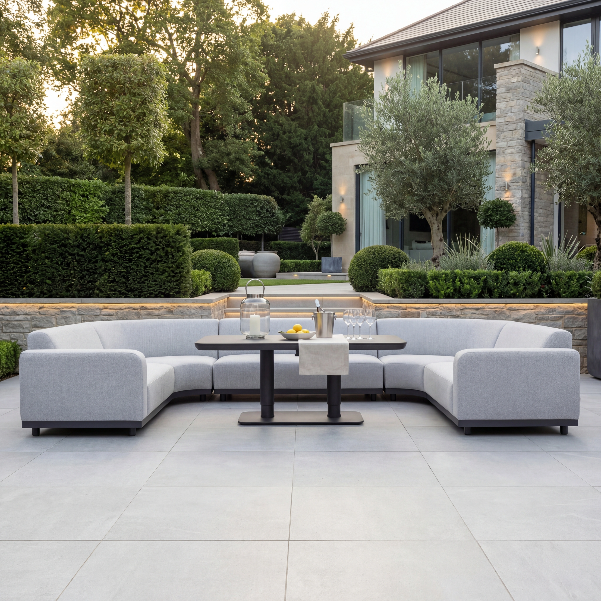 The Luna Outdoor Fabric Curved U-Shape Sofa Set with Altura Rising Table in Oyster Grey adds style and durable, all-weather comfort to a patio, featuring solution-dyed acrylic in a landscaped garden by a modern home.