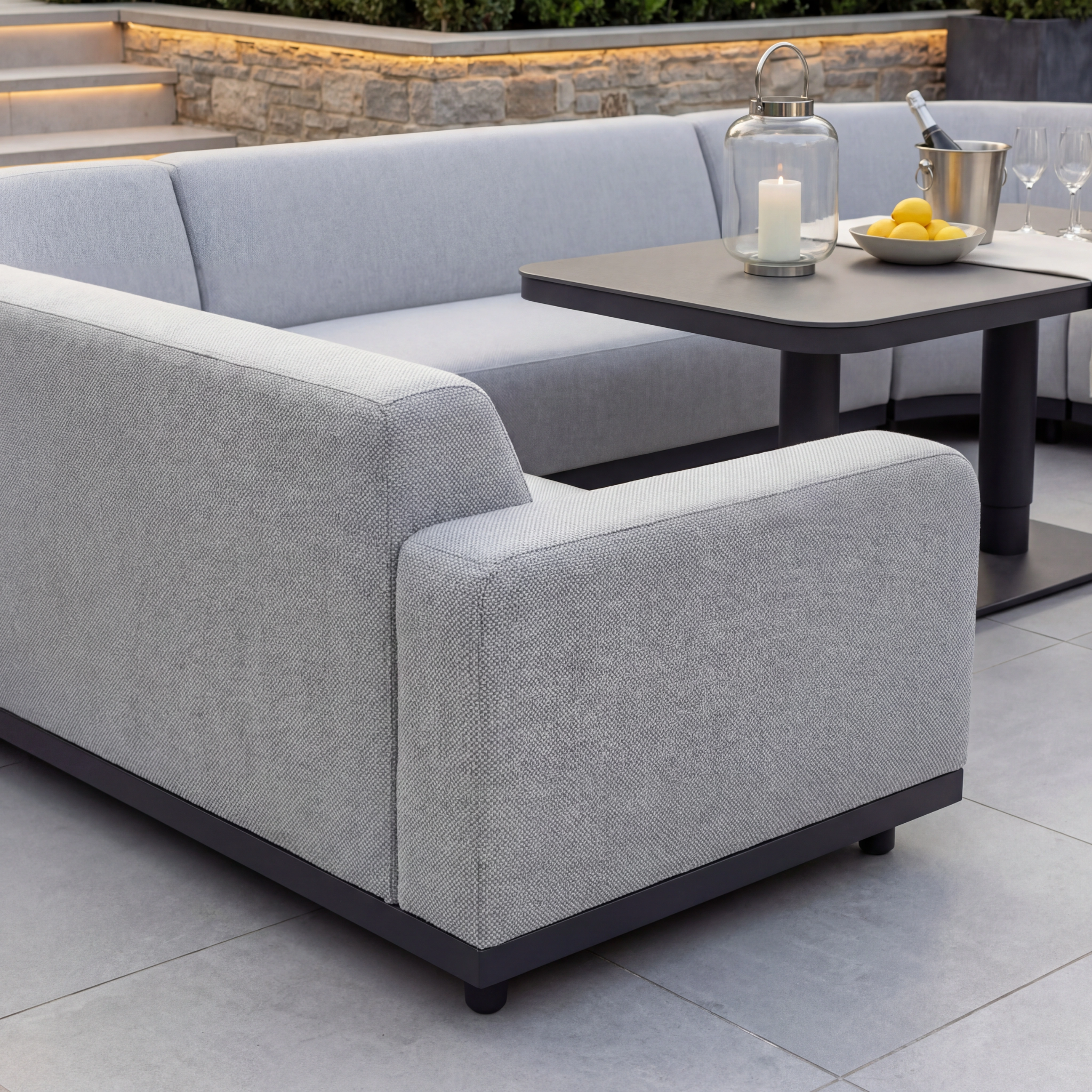 Create a modern outdoor space with the Luna Outdoor Fabric Curved U-Shape Sofa Set and Altura Rising Table in Oyster Grey, paired with candles, lanterns, lemons, and wine glasses for stylish all-weather entertaining.