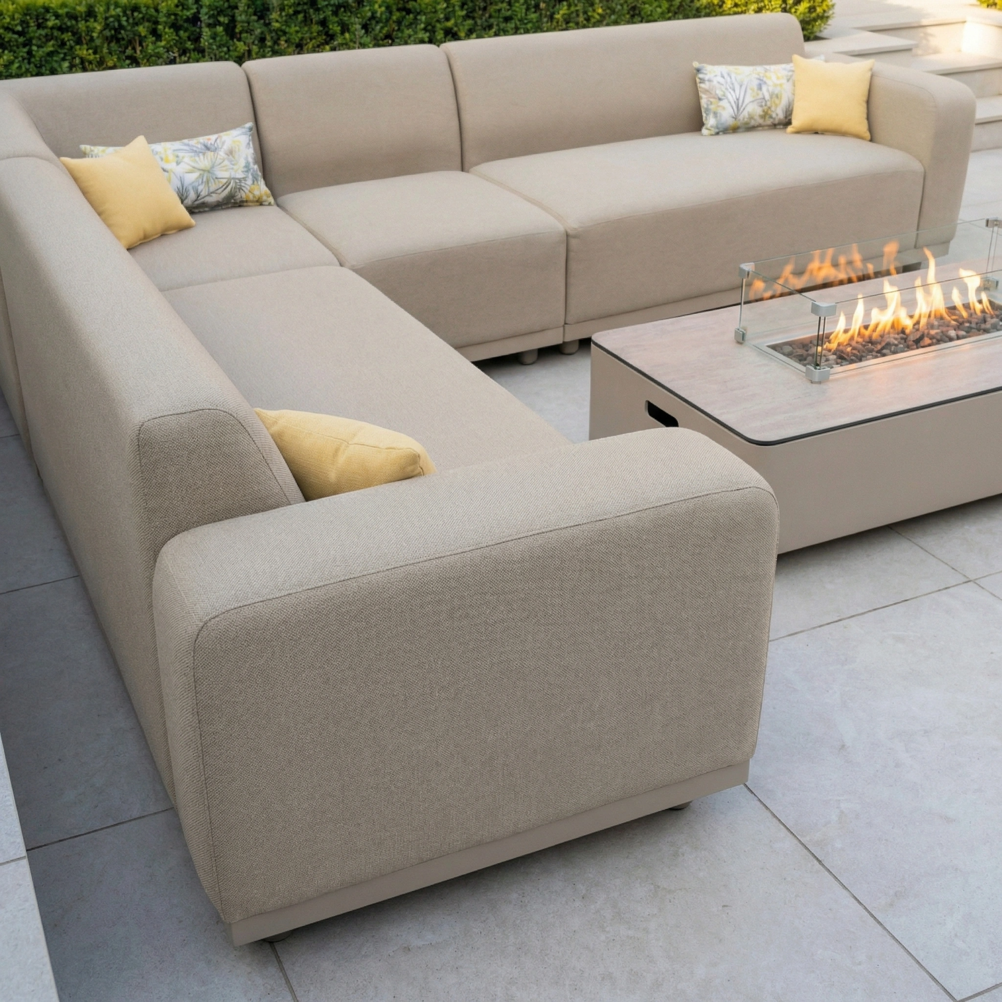 The Luna Outdoor Fabric Large Corner Group Set in Fawn, featuring yellow pillows and all-weather fabric, sits beside a modern firepit coffee table on a tiled patio.