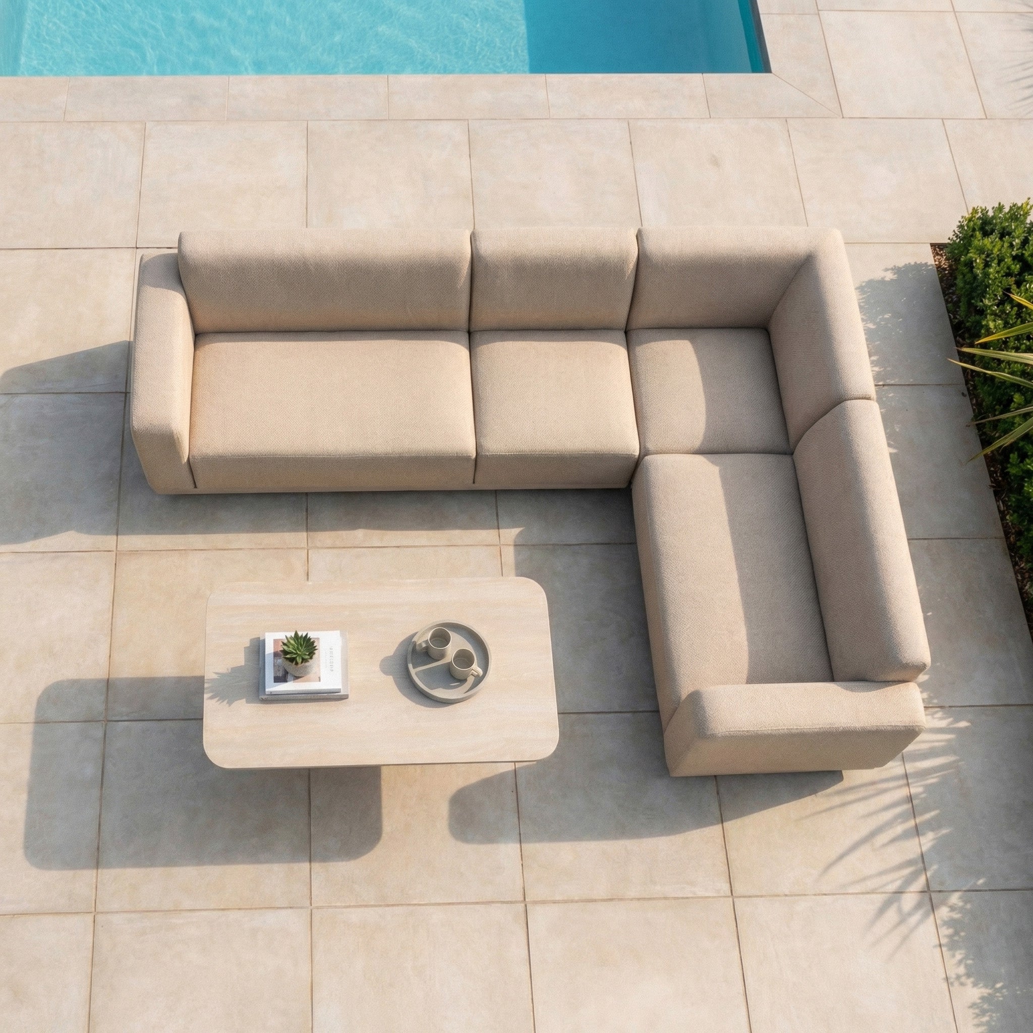 The Luna Outdoor Fabric Large Corner Sofa Set with Altura Rising Table in Fawn features a beige L-shaped design and stain-resistant fabric, shown by a pool on a tiled patio and viewed from above.