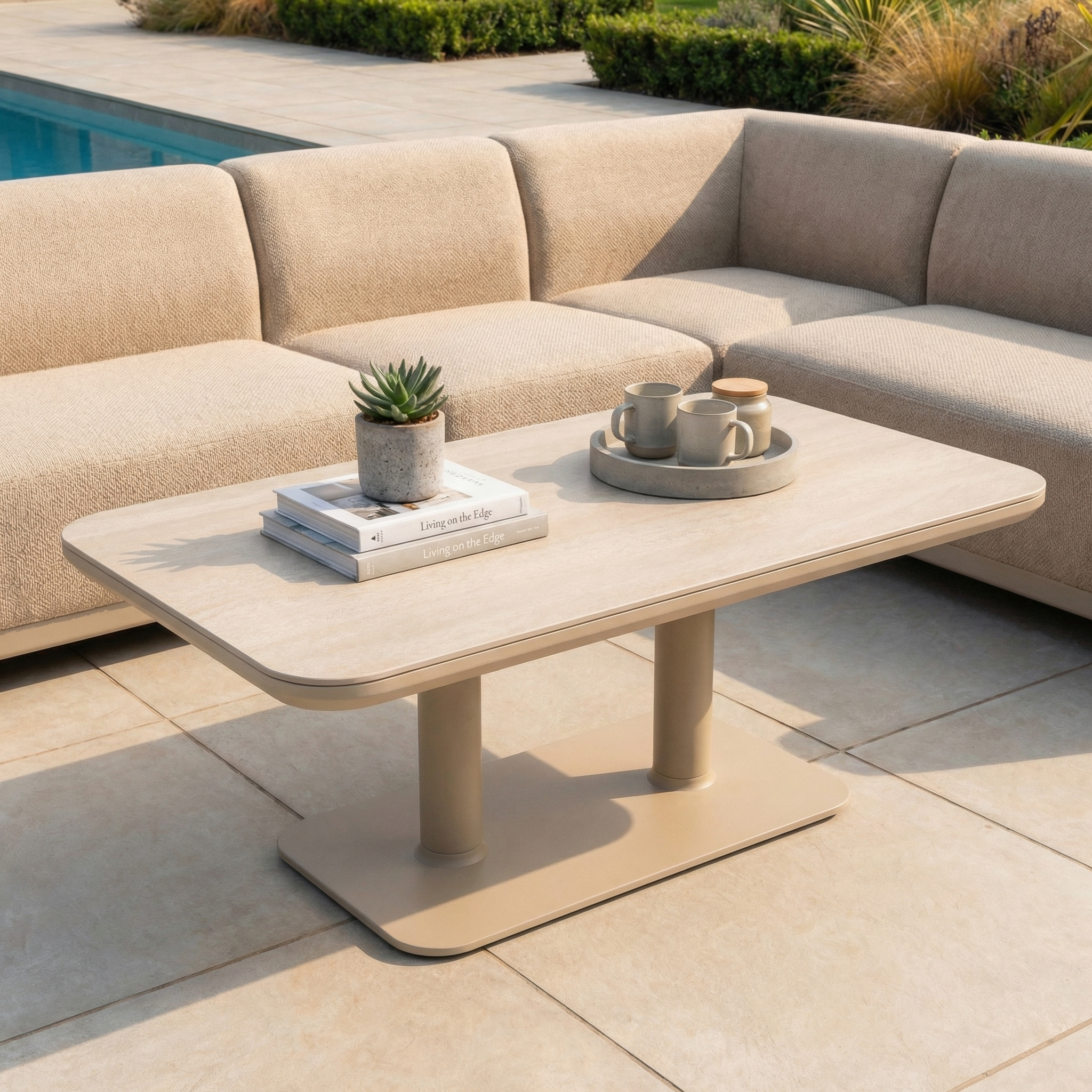 The Luna Outdoor Fabric Large Corner Sofa Set with Altura Rising Table in Fawn sits by the pool, featuring weather-resistant cushions, a tray of cups, and a potted plant on the table.