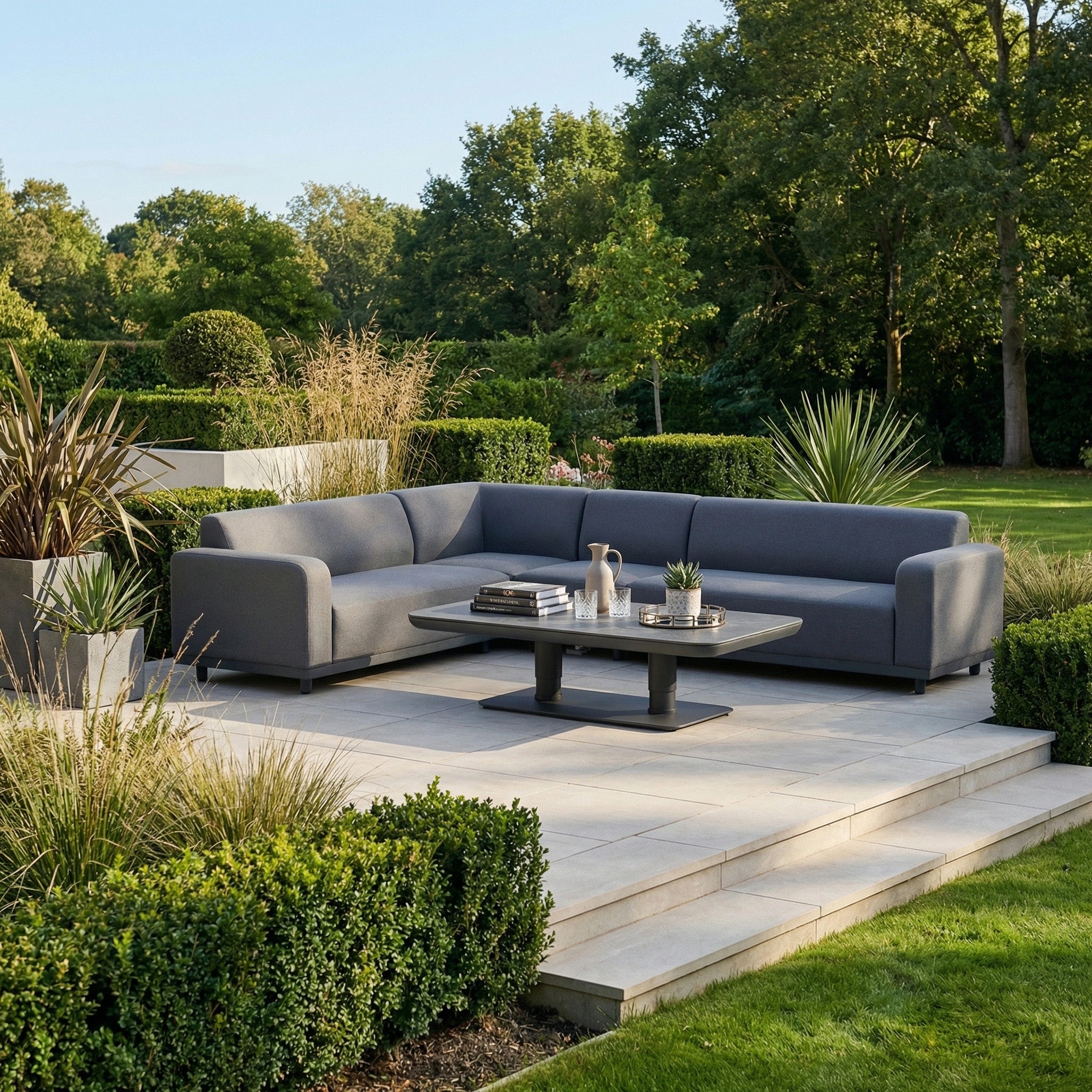 The Luna Outdoor Fabric Large Corner Sofa Set with Altura Rising Table in Grey sits on a tiled patio, boasting stain-resistant fabric and is surrounded by a lush green garden and trees.