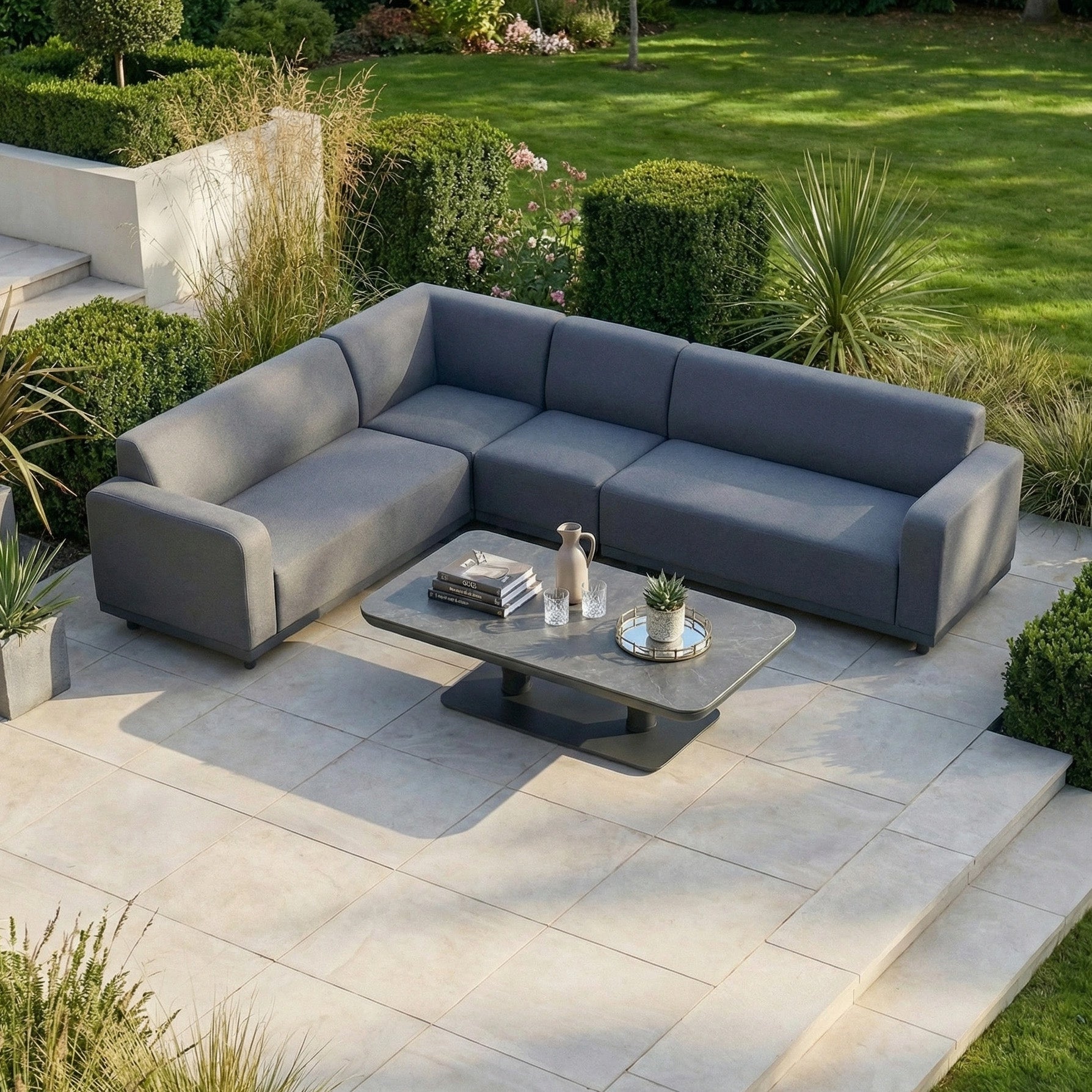 The Luna Outdoor Fabric Large Corner Sofa Set with Altura Rising Table in Grey sits on a tiled patio with stain-resistant fabric, surrounded by green plants and grass.