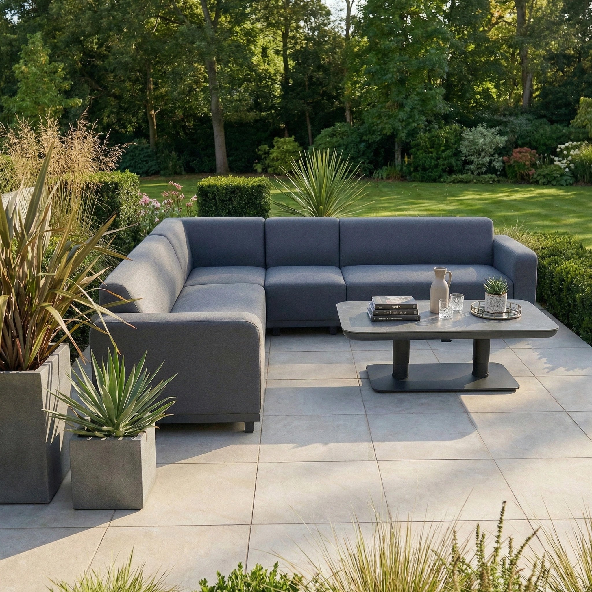 The Luna Outdoor Fabric Large Corner Sofa Set with Altura Rising Table in Grey sits on a tiled patio, surrounded by lush greenery, offering stylish, stain-resistant comfort for your outdoor space.