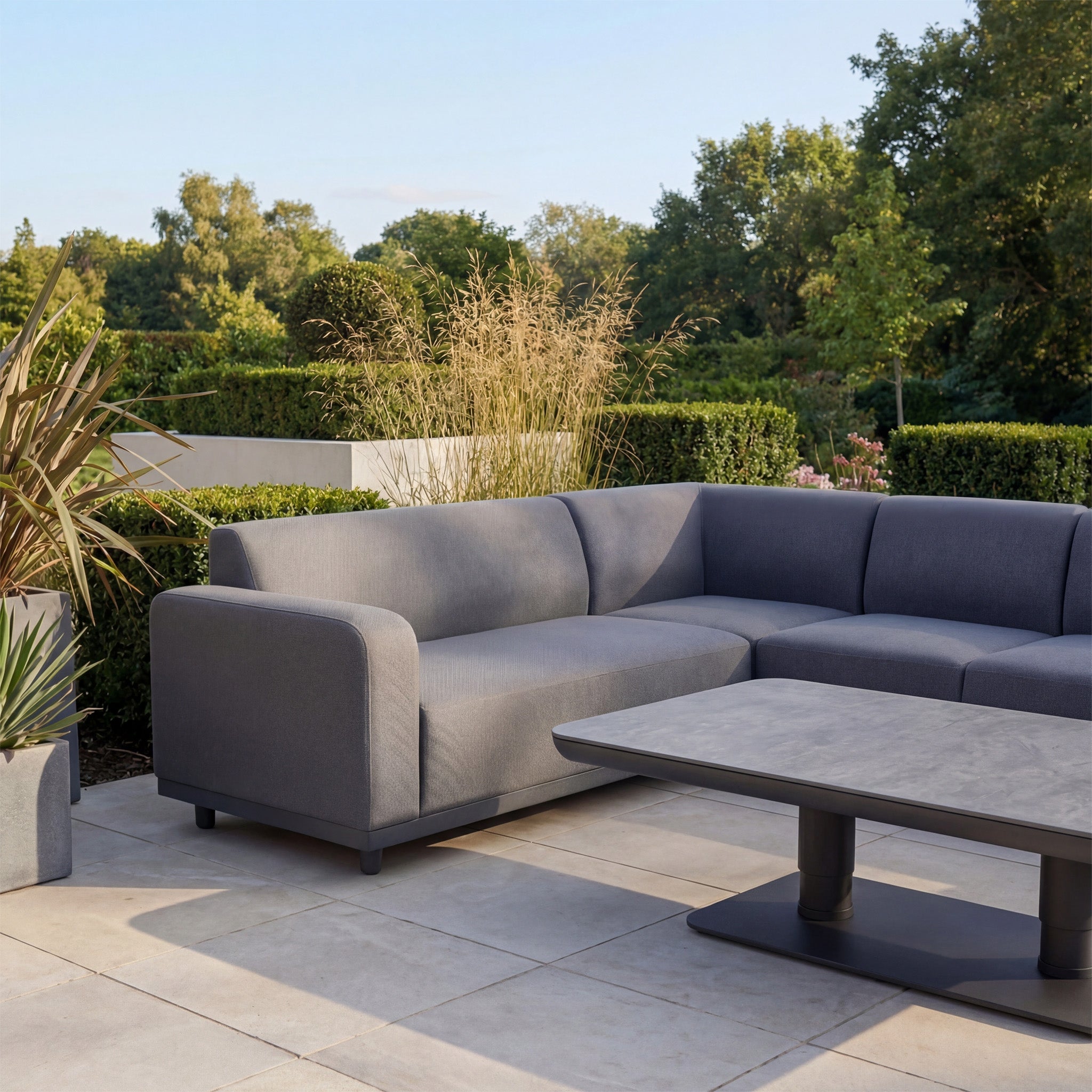 The Luna Outdoor Fabric Large Corner Sofa Set with Altura Rising Table in Grey features stain-resistant fabric and a rectangular table, perfect for tiled patios surrounded by greenery and plants.