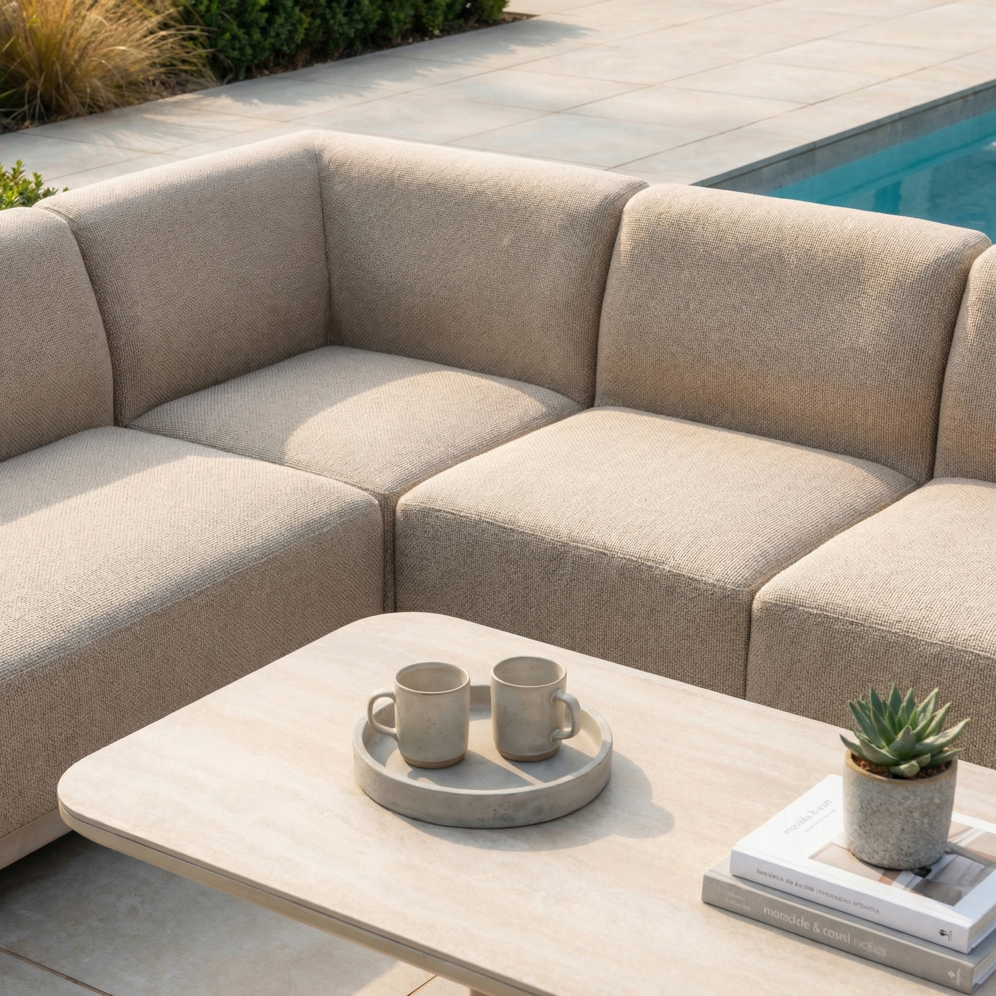The Luna Outdoor Fabric Large Corner Sofa Set with Altura Rising Table in Fawn, crafted from weather-resistant fabric, sits by the pool. The rising table holds two mugs, books, and a small potted succulent.