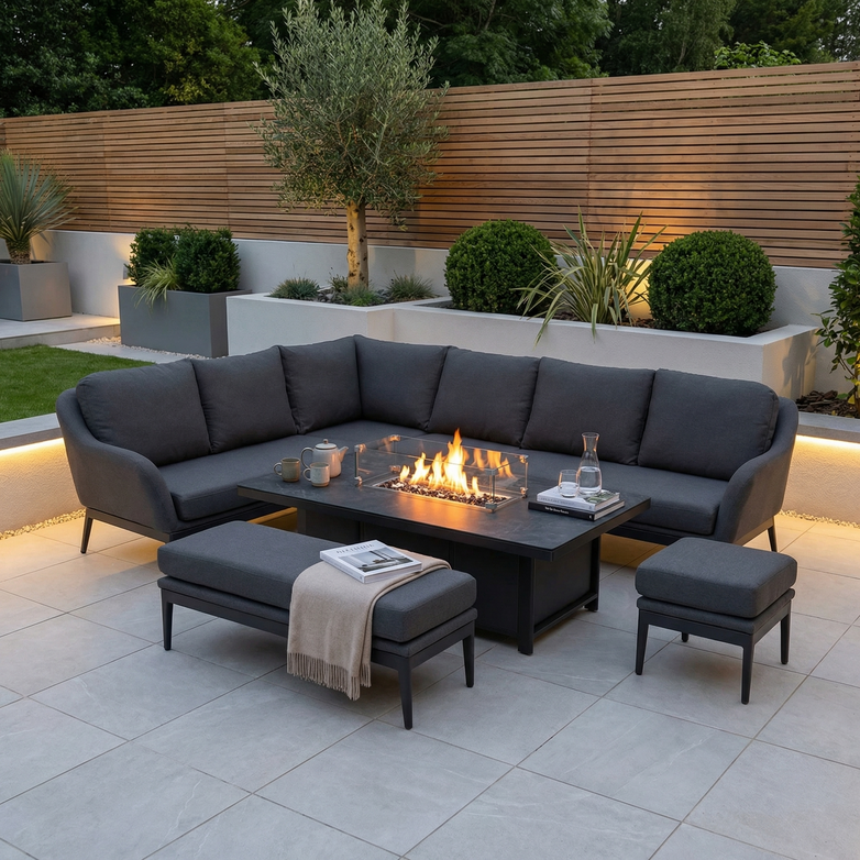 Modern patio featuring the Luna Outdoor Fabric Rectangular Corner Dining Set with Rising Firepit Table in Grey (Left Hand), surrounded by greenery at dusk.