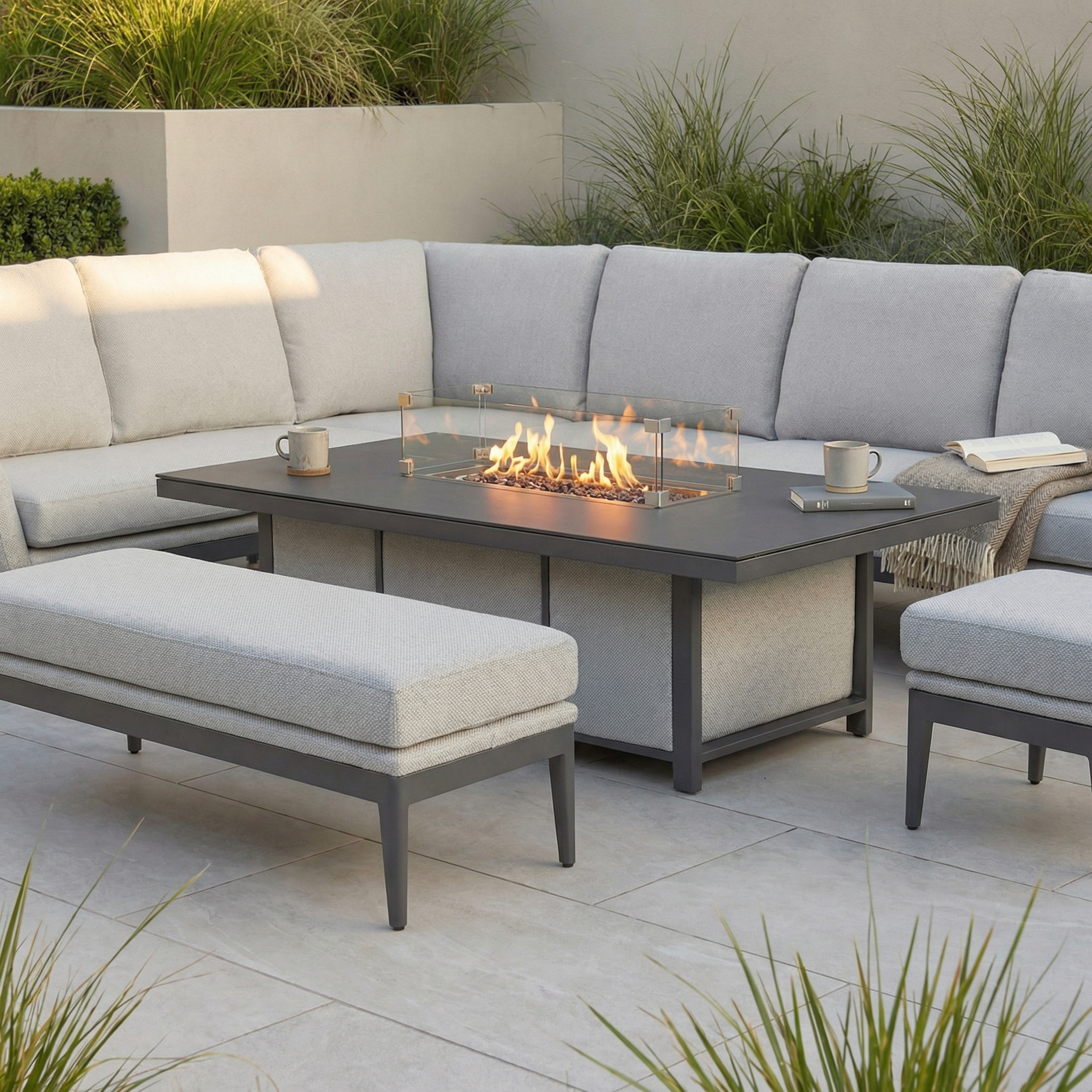 The Luna Outdoor Fabric Rectangular Corner Dining Set with Rising Firepit Table in Oyster Grey (Left Hand) and matching bench creates a relaxing patio oasis surrounded by greenery with maintenance-free garden furniture.