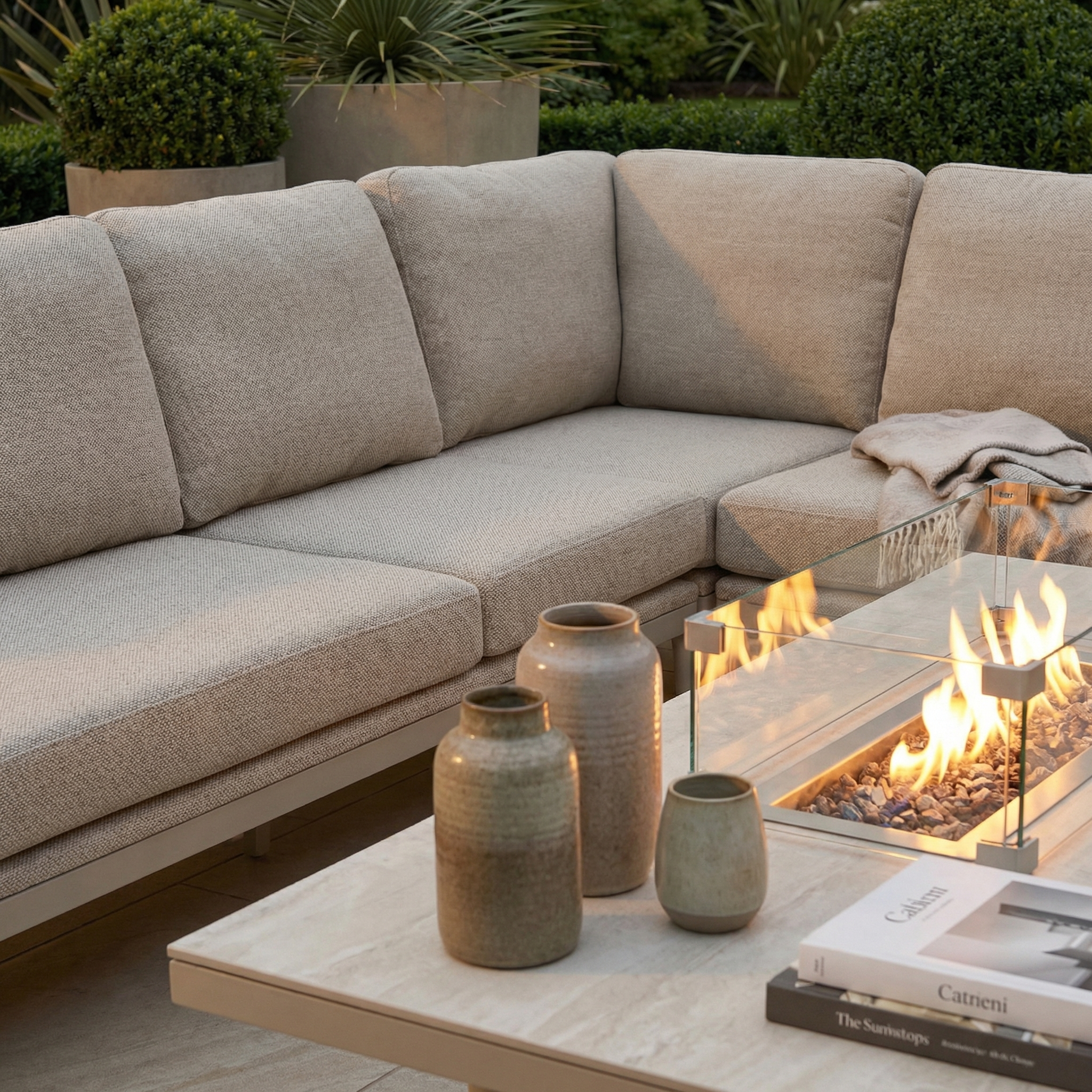 The Luna Outdoor Fabric Rectangular Corner Dining Set in Fawn (Right Hand) features modern beige seating, a cozy blanket, rising firepit table, ceramic vases, and books—perfect for stylish patio lounging.