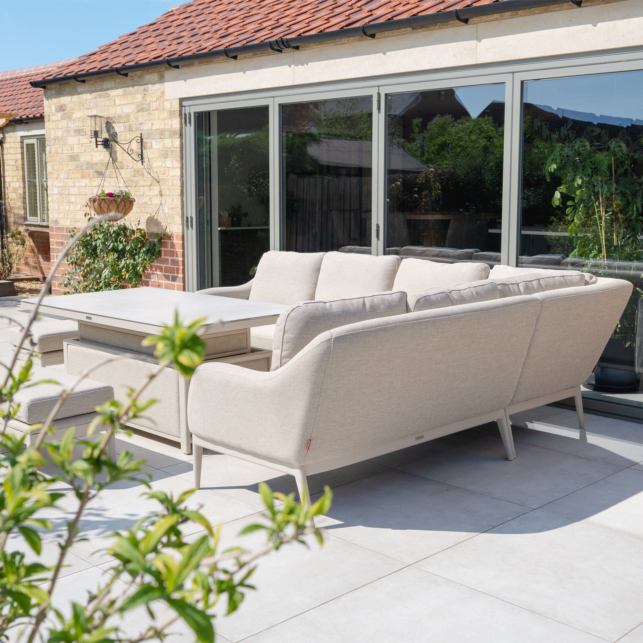 The Luna Outdoor Fabric Rectangular Corner Dining Set with Rising Table in Fawn (Left Hand) features a maintenance-free beige fabric, a glass rising table, and is perfect for sunny patios near sliding doors and lush plants.