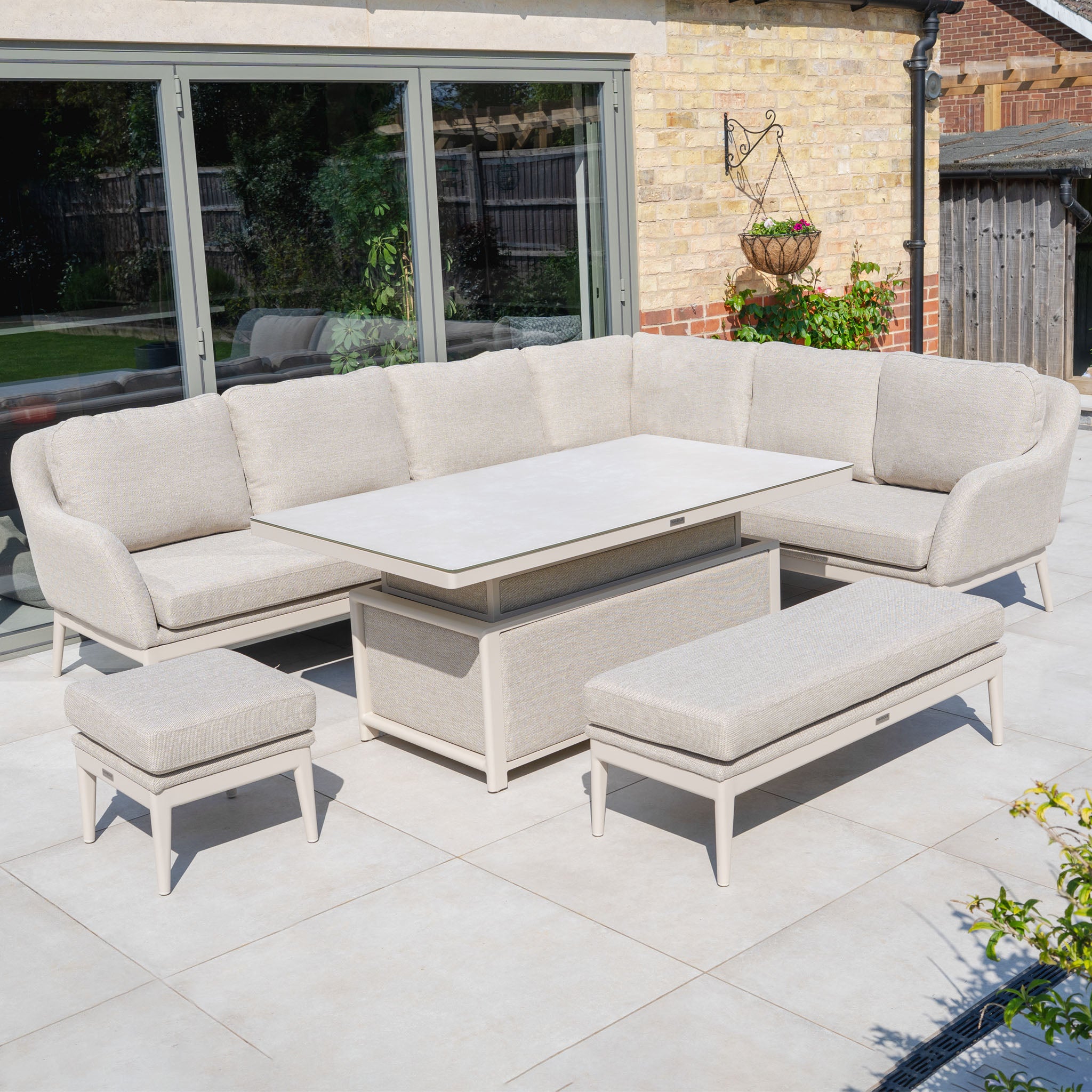 The Luna Outdoor Fabric Rectangular Corner Dining Set with Rising Table in Fawn (Left Hand) includes a sofa, bench, stool, and table on a tiled patio near sliding glass doors.