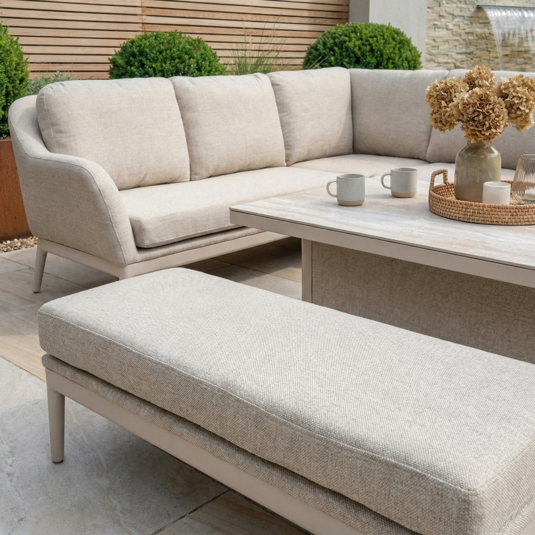 Luna Outdoor Fabric Rectangular Corner Dining Set with Rising Table in Fawn (Left Hand), featuring a bench, coffee table, and vase on a tiled patio with plants; crafted from all-weather material for lasting style.