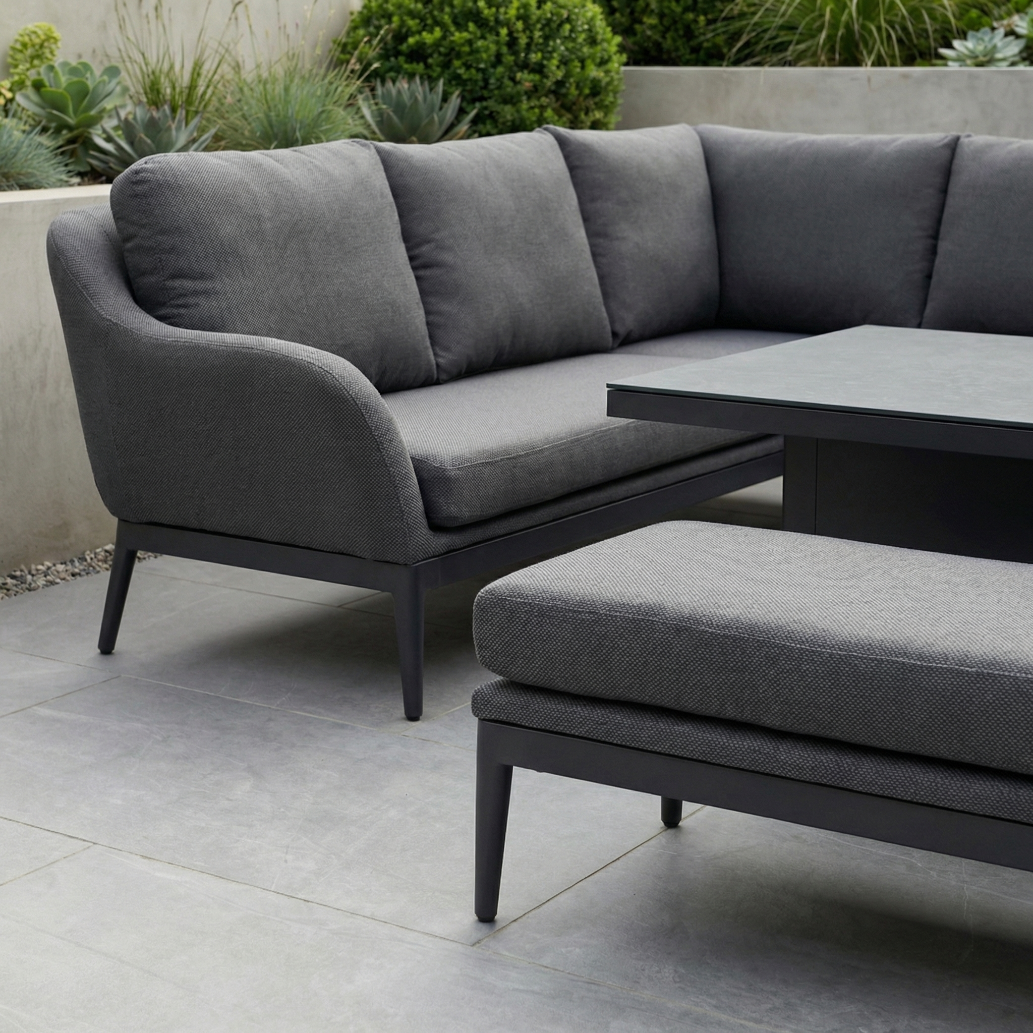 Luna Outdoor Fabric Rectangular Corner Dining Set with Rising Table in Grey (Left Hand) on a tiled patio, featuring dark gray cushions and plants in the background.