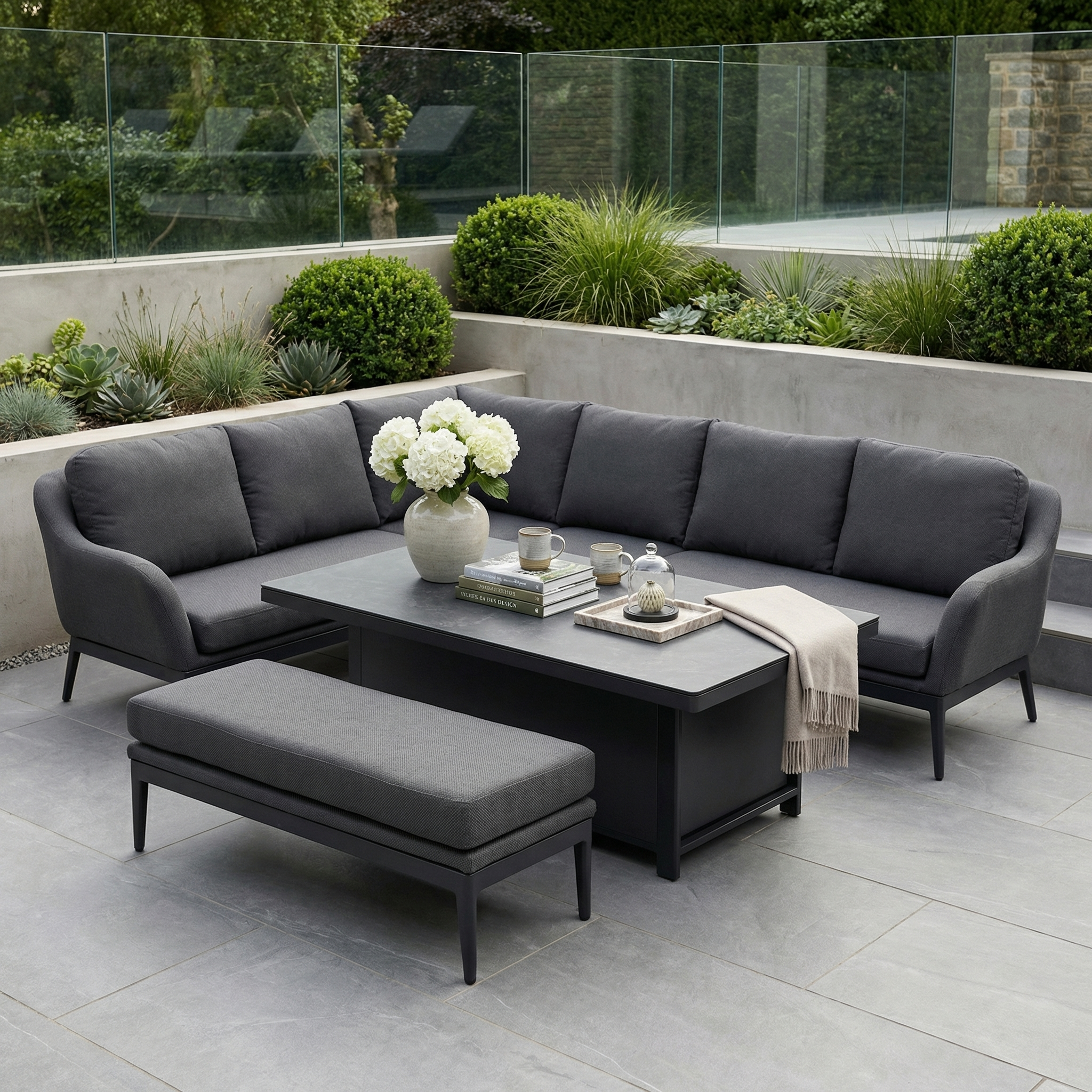 The Luna Outdoor Fabric Rectangular Corner Dining Set with Rising Table in Grey (Left Hand) features comfy gray cushions and a versatile rising table, perfect for dining or relaxing outdoors amid lush greenery.