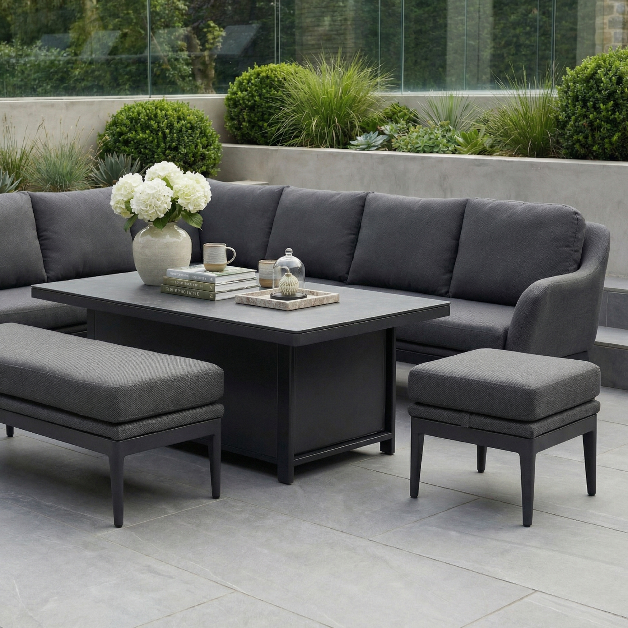 Modern outdoor patio featuring the Luna Outdoor Fabric Rectangular Corner Dining Set with Rising Table in Grey (Left Hand), benches, a coffee table, and potted plants in the background.