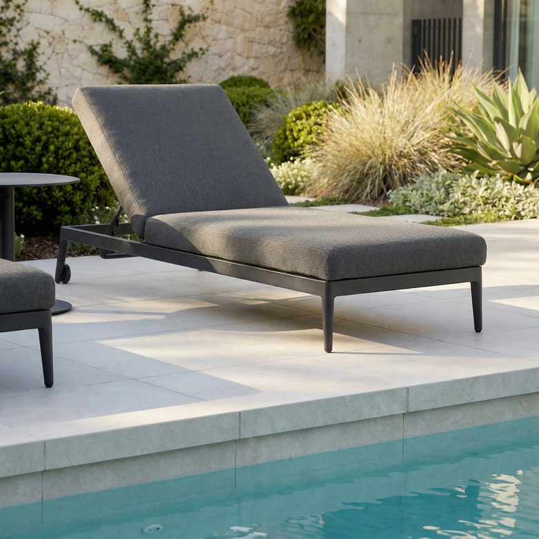The Luna Outdoor Fabric Sun Lounger in Grey sits poolside, crafted from all-weather material and set amid lush greenery and modern landscaping.