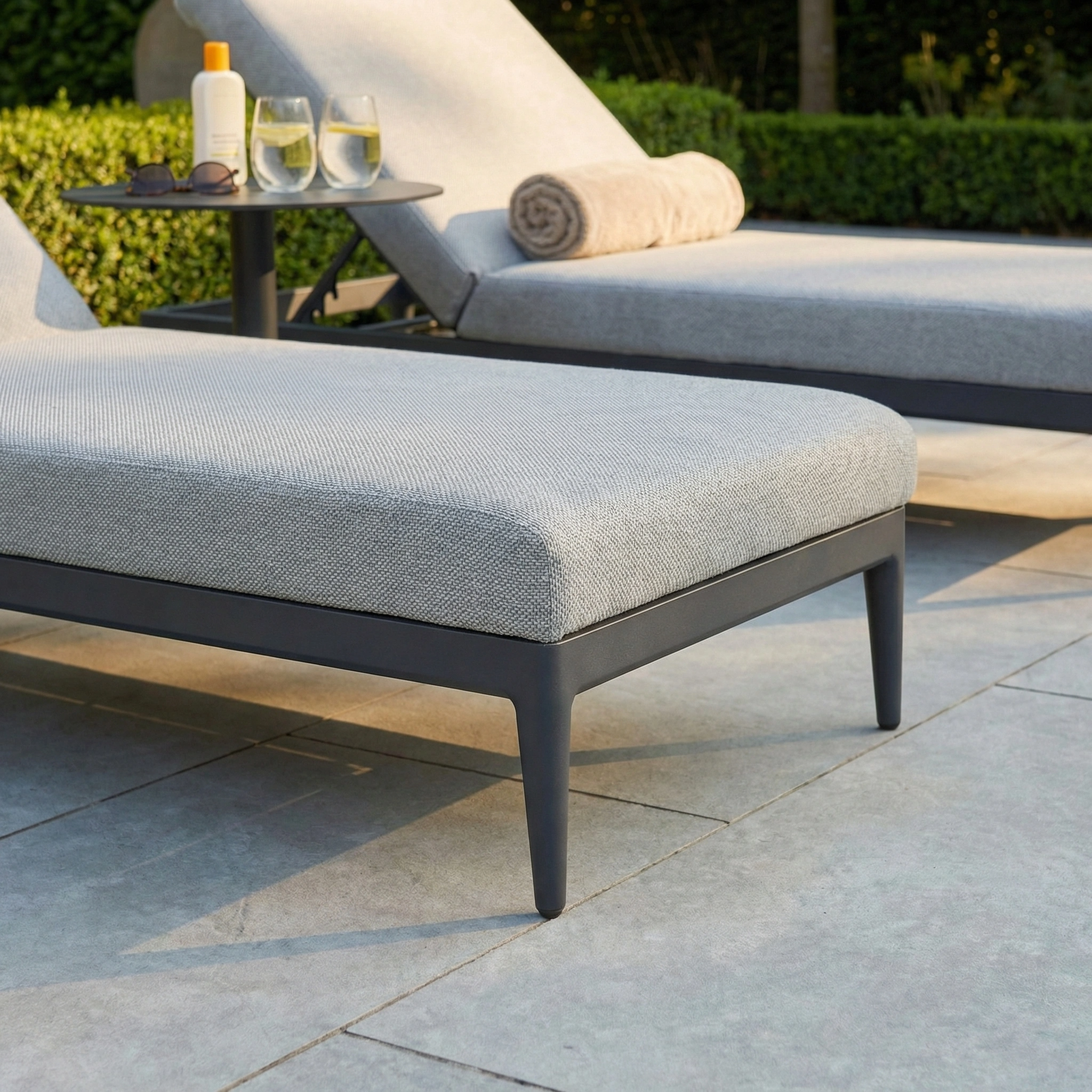 Two Luna Outdoor Fabric Sun Loungers in Oyster Grey, crafted from all-weather material, are set by the pool alongside drinks, a towel, and sunscreen on a side table—ideal for relaxing outdoors in style and comfort.