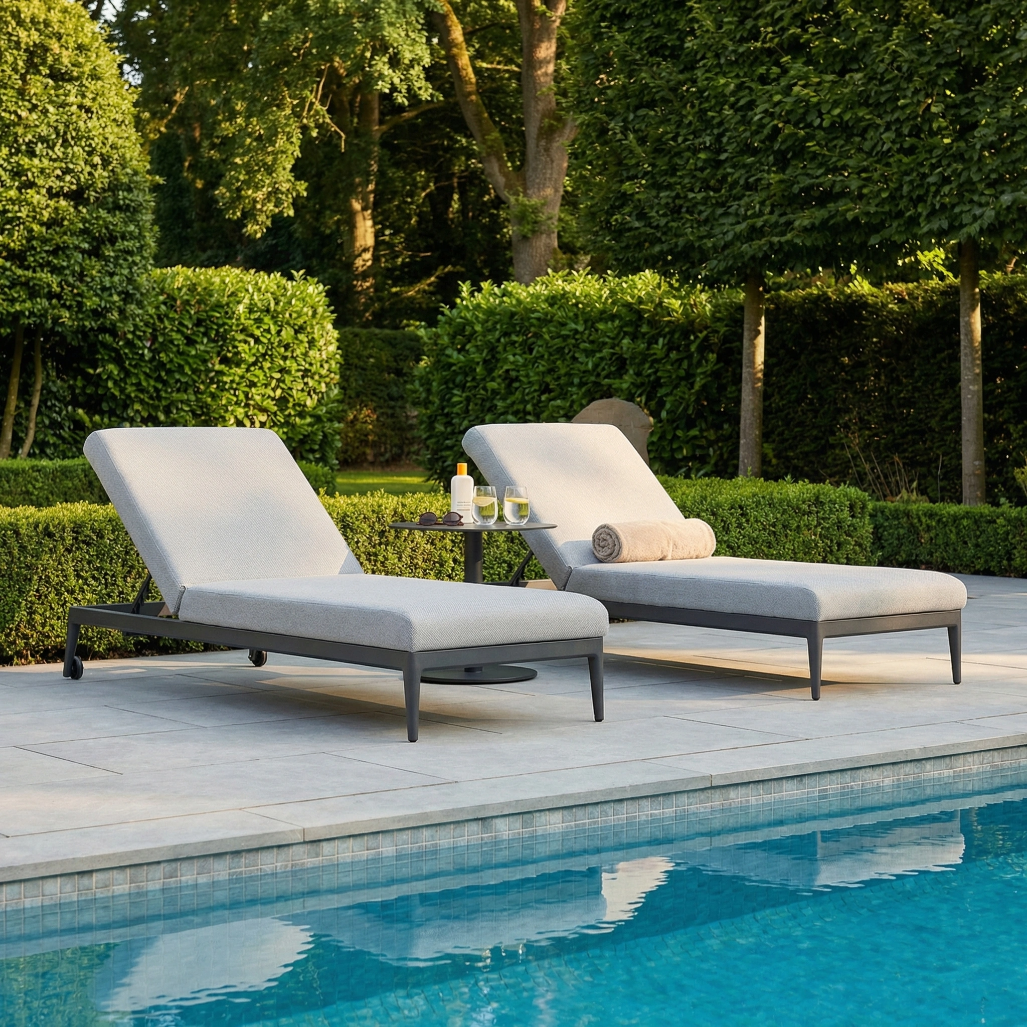 Two Luna Outdoor Fabric Sun Loungers in Oyster Grey, along with a small table holding drinks and a towel, are placed poolside amid lush greenery.