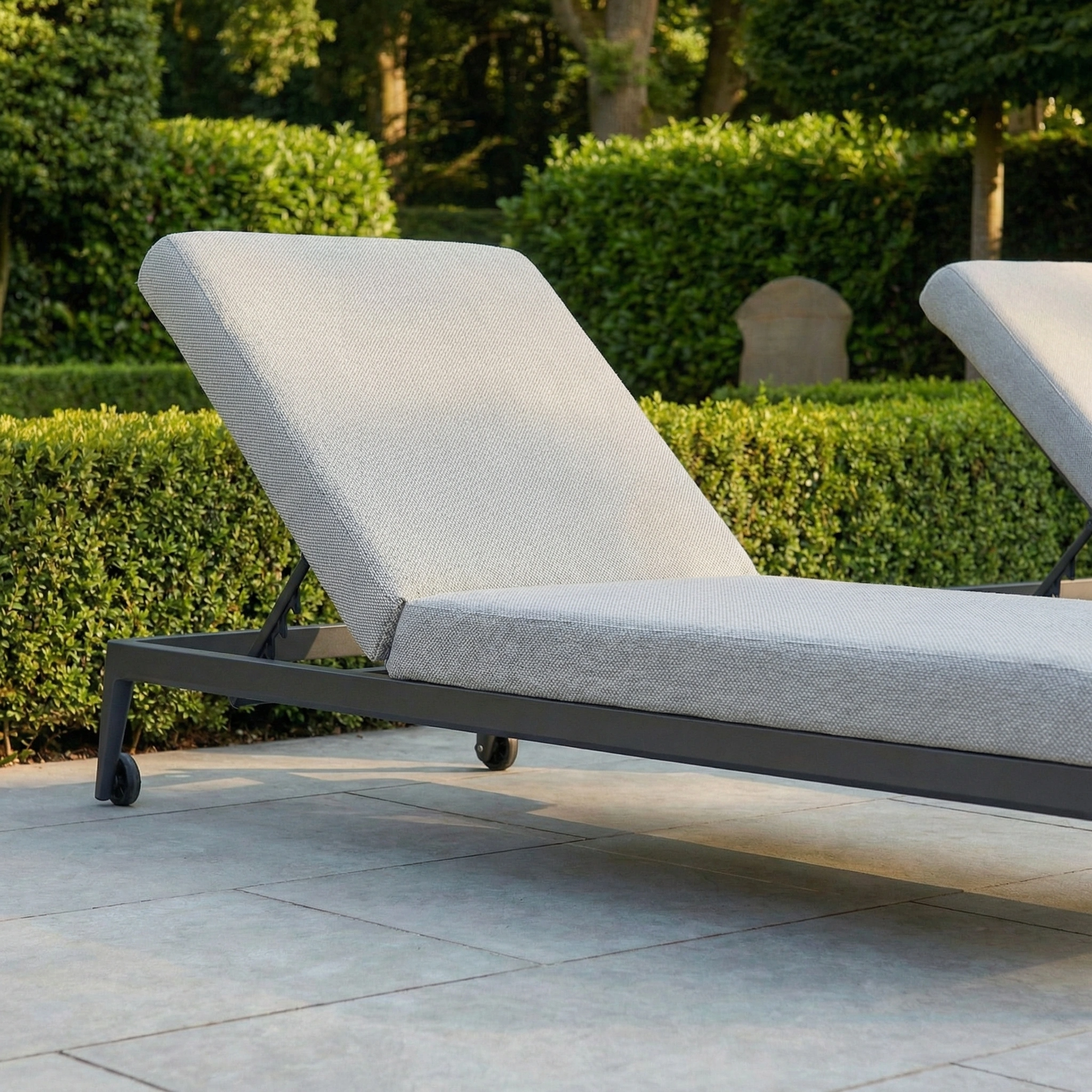 The Luna Outdoor Fabric Sun Lounger in Oyster Grey rests on a tiled patio, framed by lush green hedges and trees.